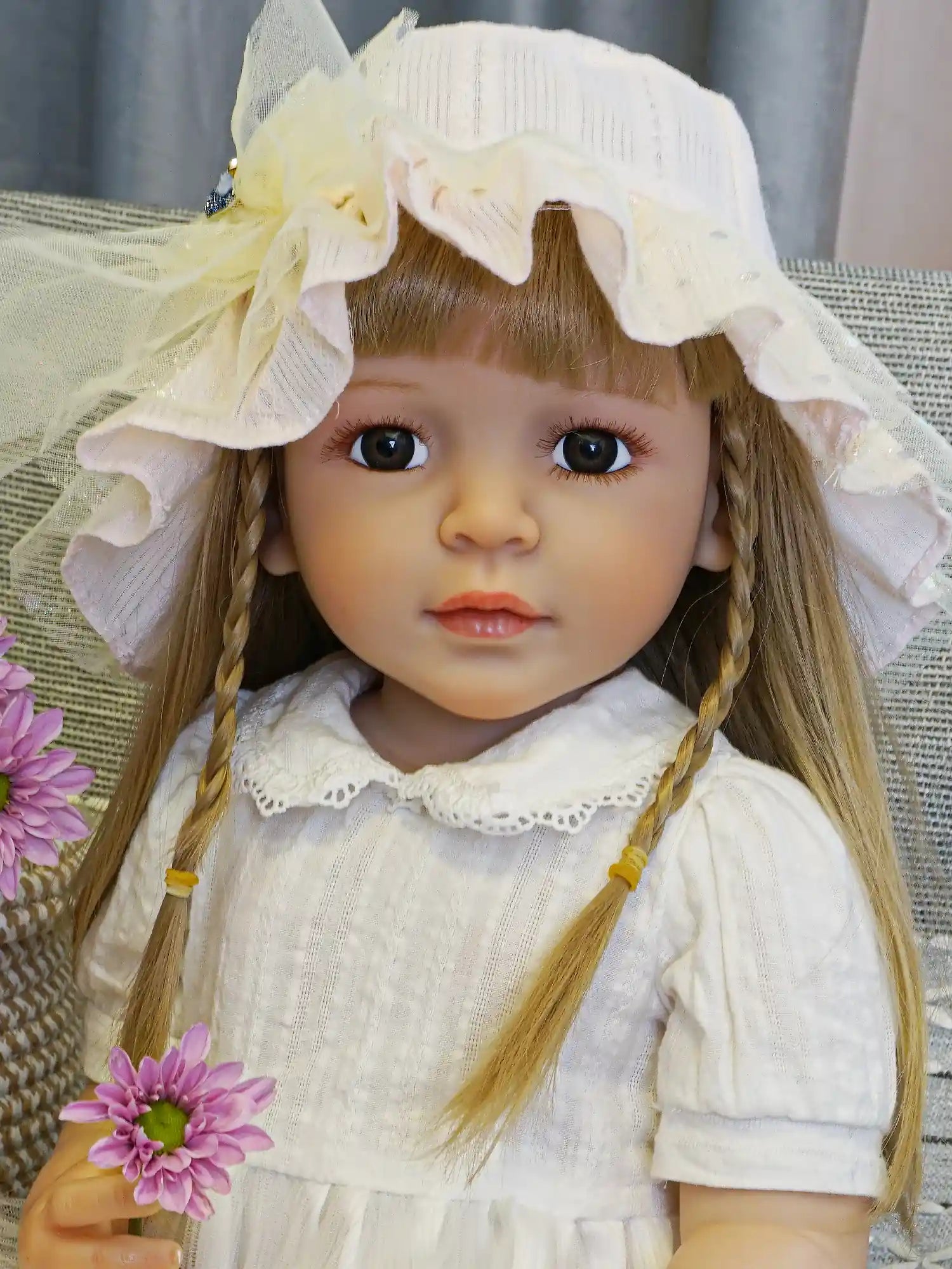 Lifelike reborn doll in a white lace dress and yellow sunhat, seated on a cushion.