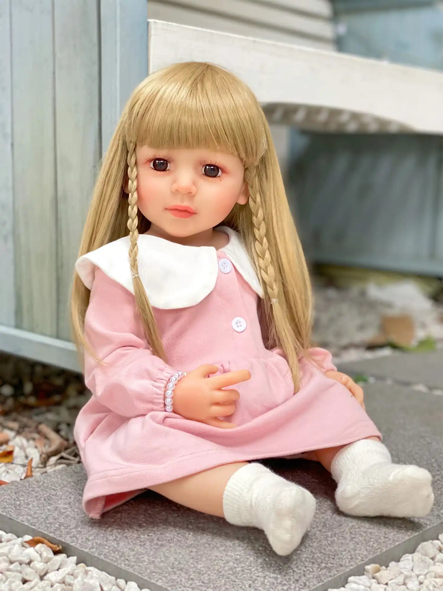 Lifelike doll with pigtails, wearing a rose-pink dress, sitting outdoors on a grey mat.