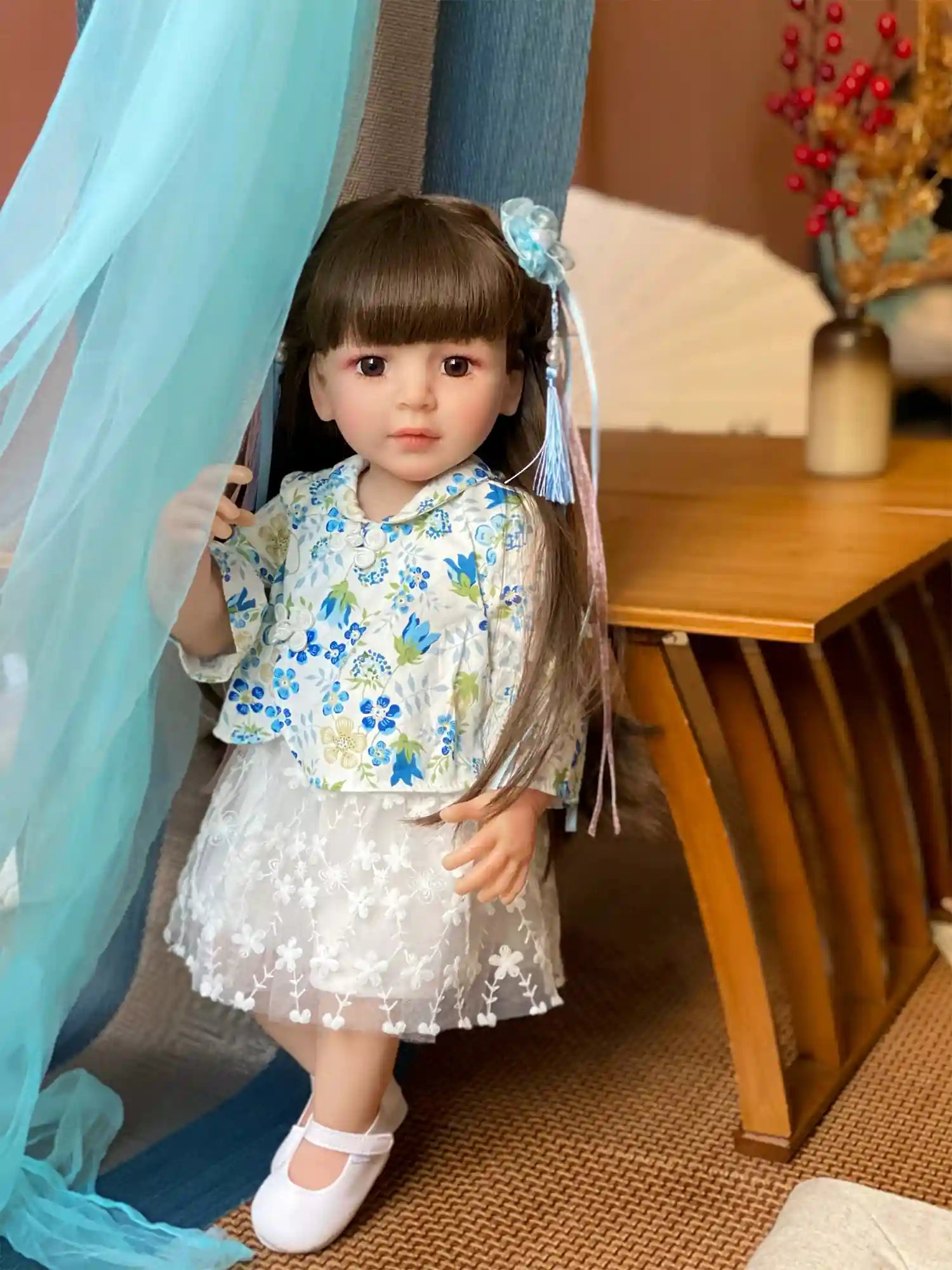 Toddler doll seated with blue floral dress and white shoes, accessorized with blue hair ribbons, against a home interior backdrop.