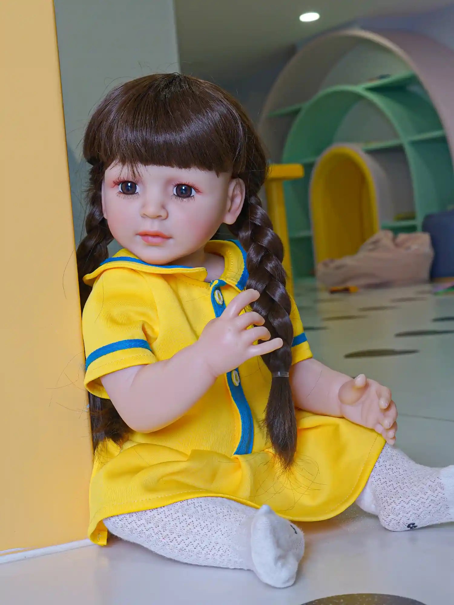 Toddler doll with braided dark hair and a blue-eyed gaze, wearing a bright yellow dress, seated in a playful nursery setting.