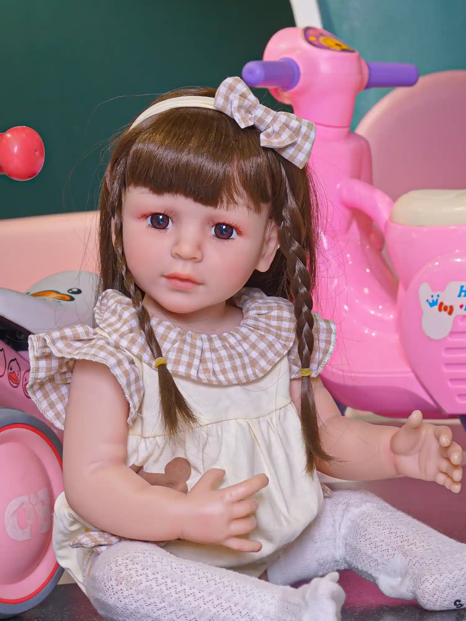 Close-up of a lifelike toddler doll with braided hair and a plaid hair bow, deep blue eyes, in a playful setting.