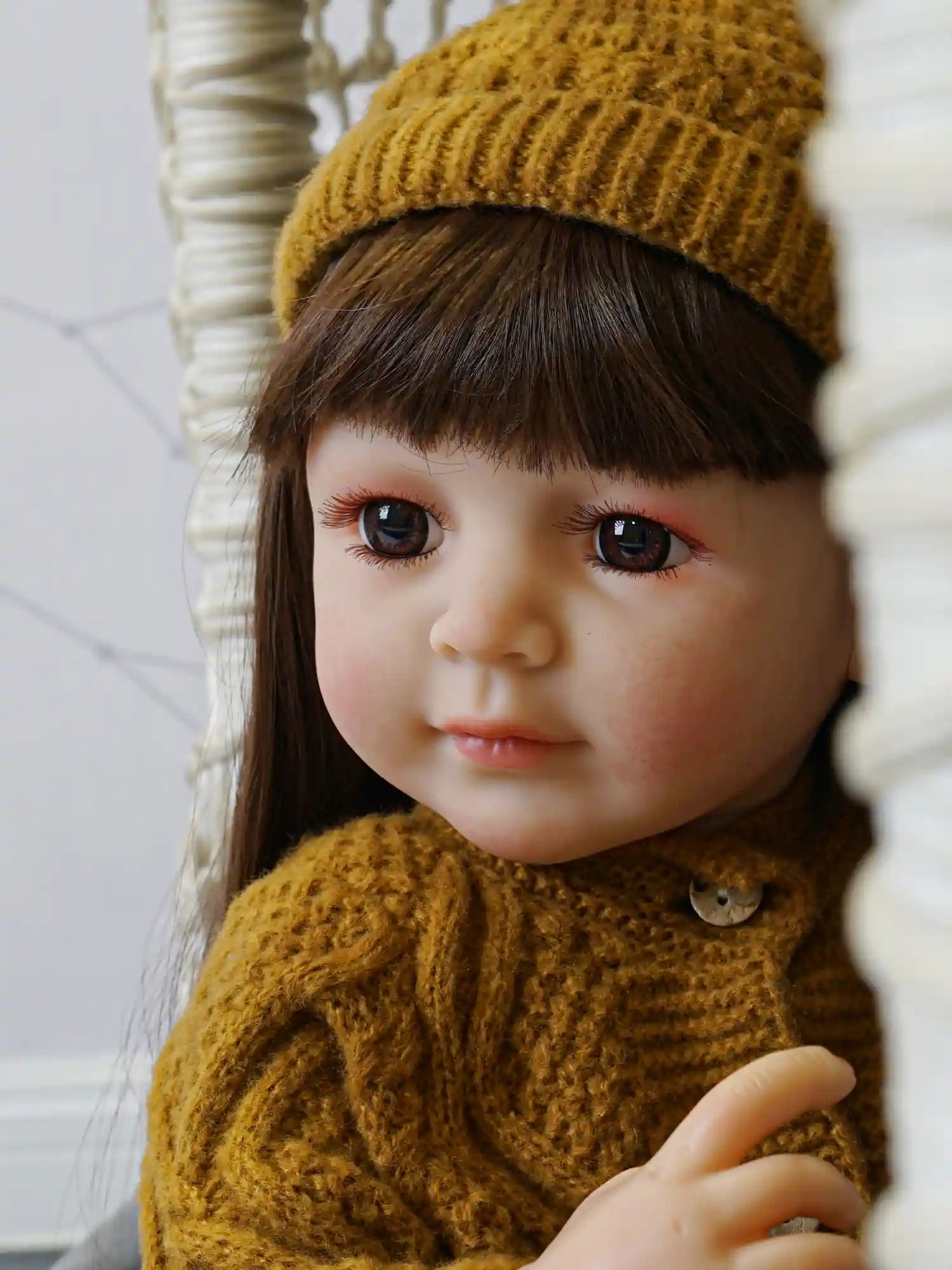 Doll with detailed features, including glossy brown hair and tender brown eyes, dressed warmly in a mustard-yellow sweater and hat, sitting against a gray patterned background.