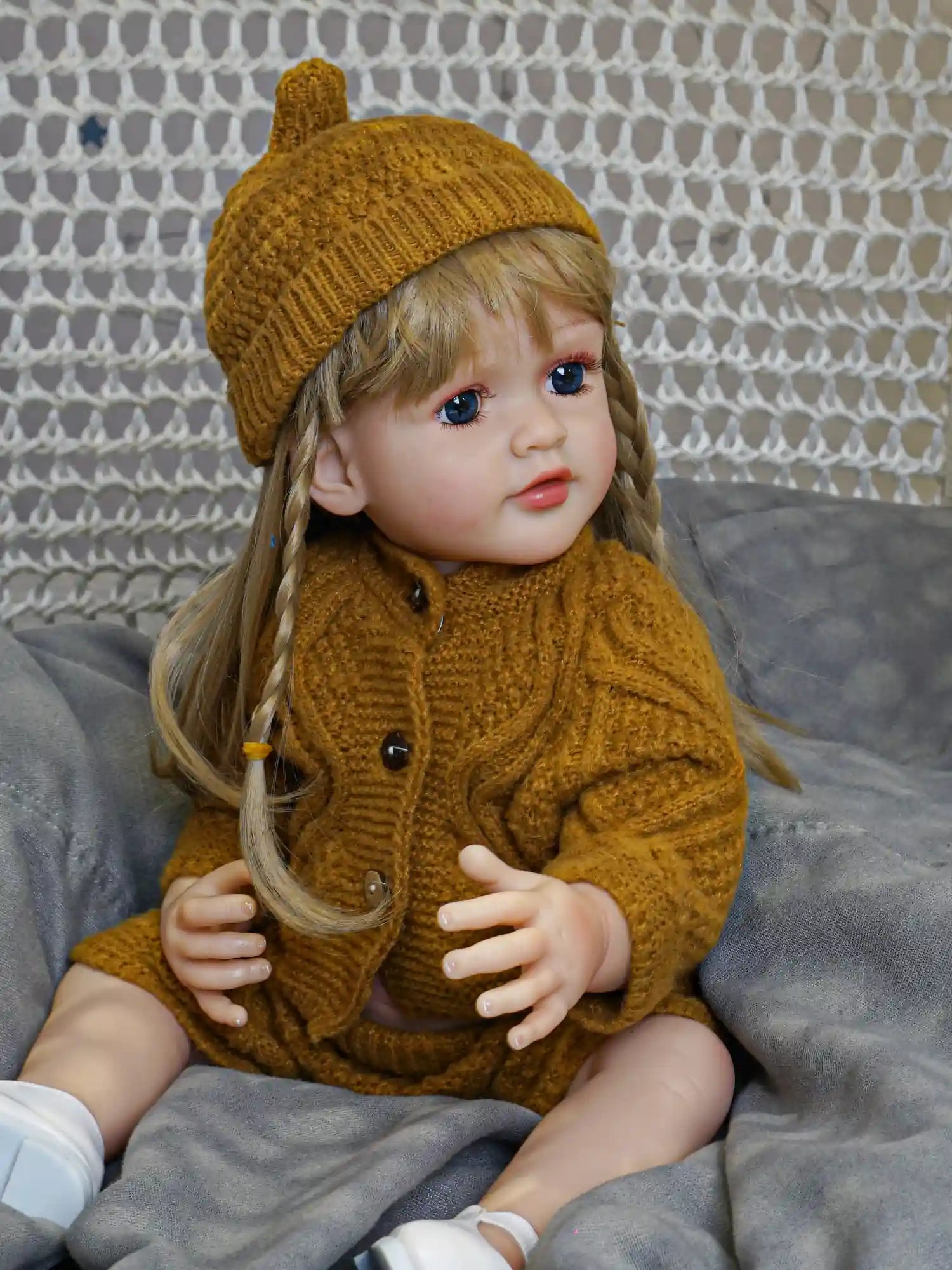 Detailed toddler doll in a relaxed seated position, featuring long blonde hair with braids and expressive blue eyes, dressed in a warm winter outfit of knitted mustard yarn.
