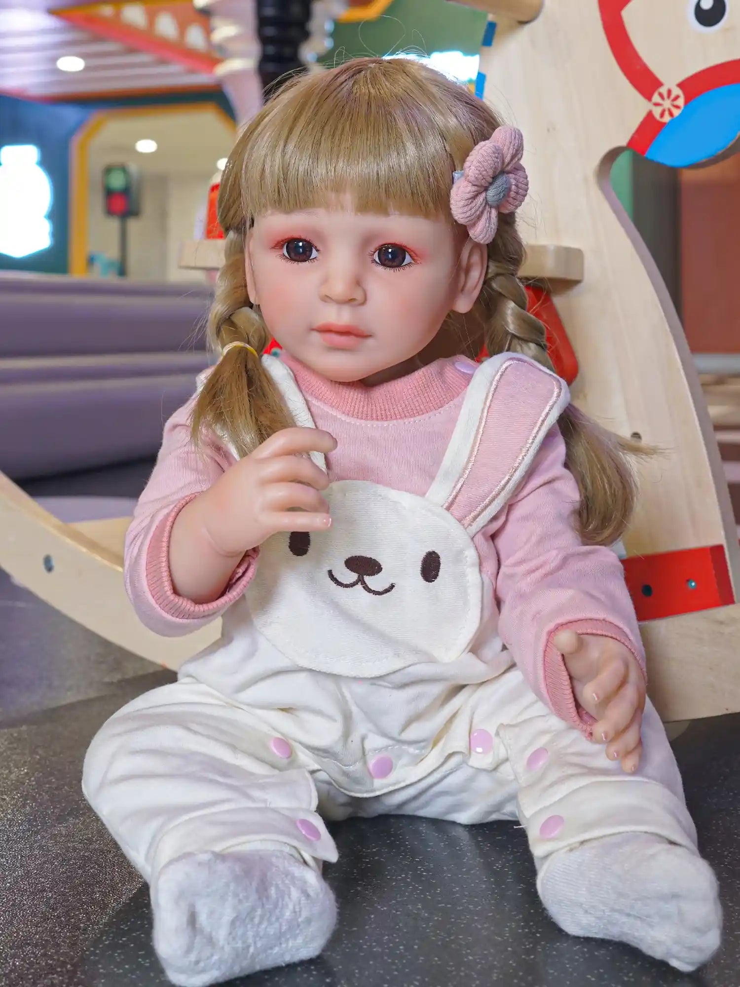 Toddler doll seated on a play mat, wearing a cozy pink and white outfit with a bear motif, in a playful indoor environment.