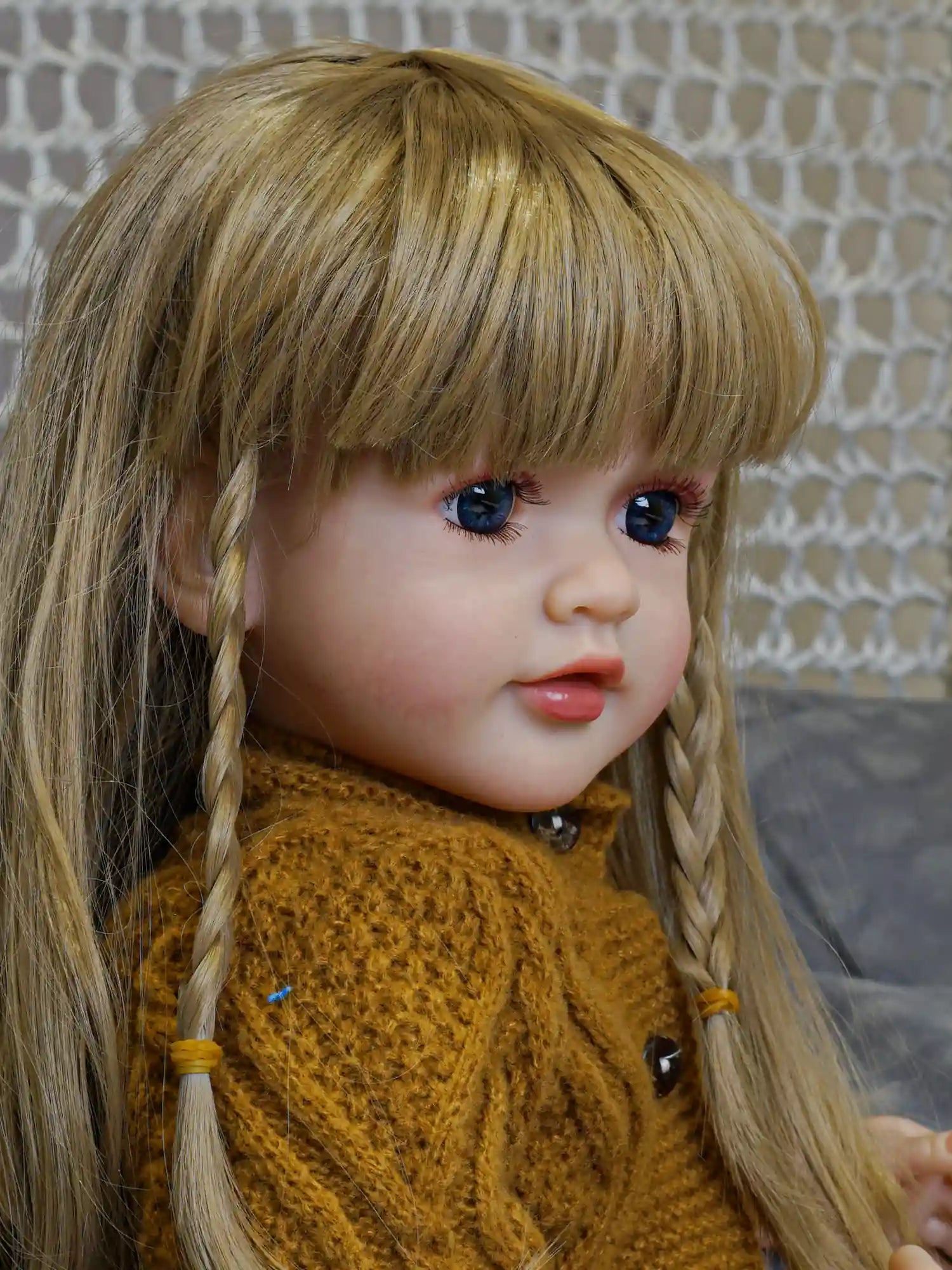 A realistic doll with braided hair and a thoughtful gaze, clothed in a chunky knit mustard sweater and hat, contrasting with the delicate grey and white background.