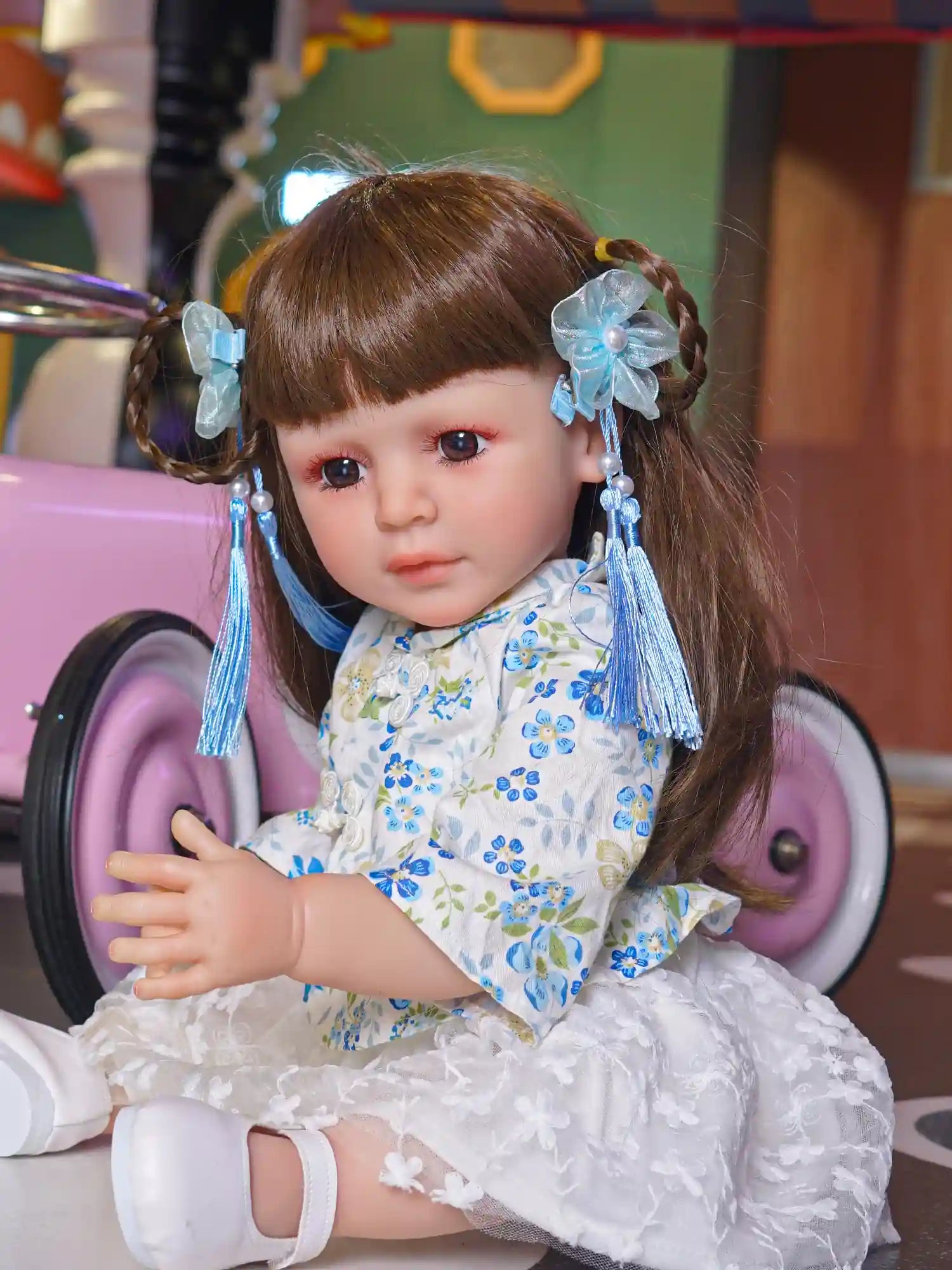 Toddler doll sitting on the floor, dressed in a blue floral dress with white lace, white shoes, and blue hair accessories, beside a pink toy car.