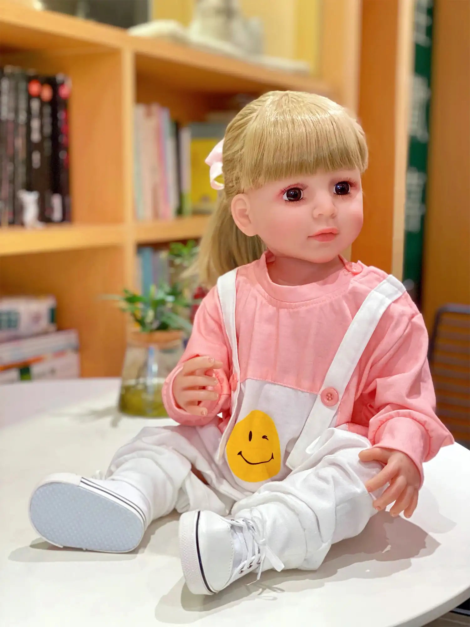 Lifelike doll with soft blonde hair and engaging blue eyes, dressed in casual play attire, in an indoor play setting.