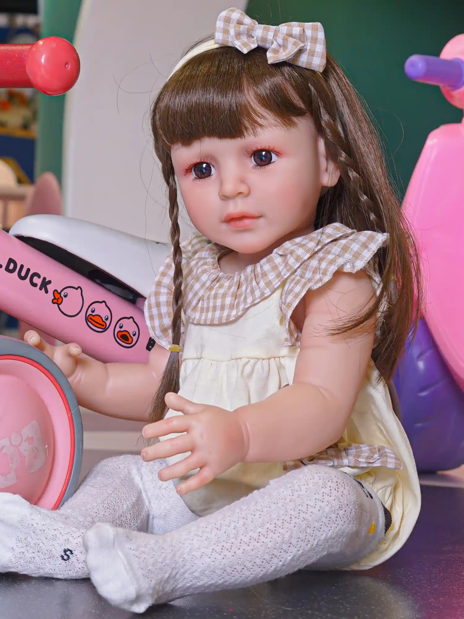 Toddler doll seated cross-legged, in a cream pinafore and white tights, with a red and pink scooter nearby.