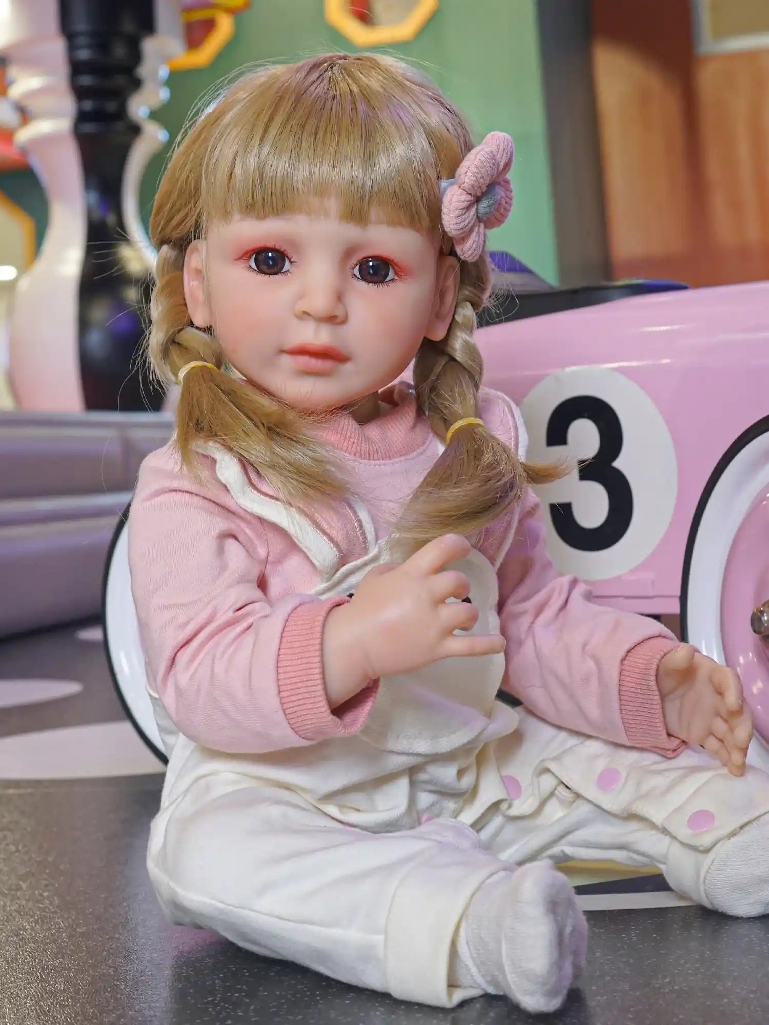 Close-up of a toddler doll with blonde braids and purple hair ties, pink cheeks, seated indoors.