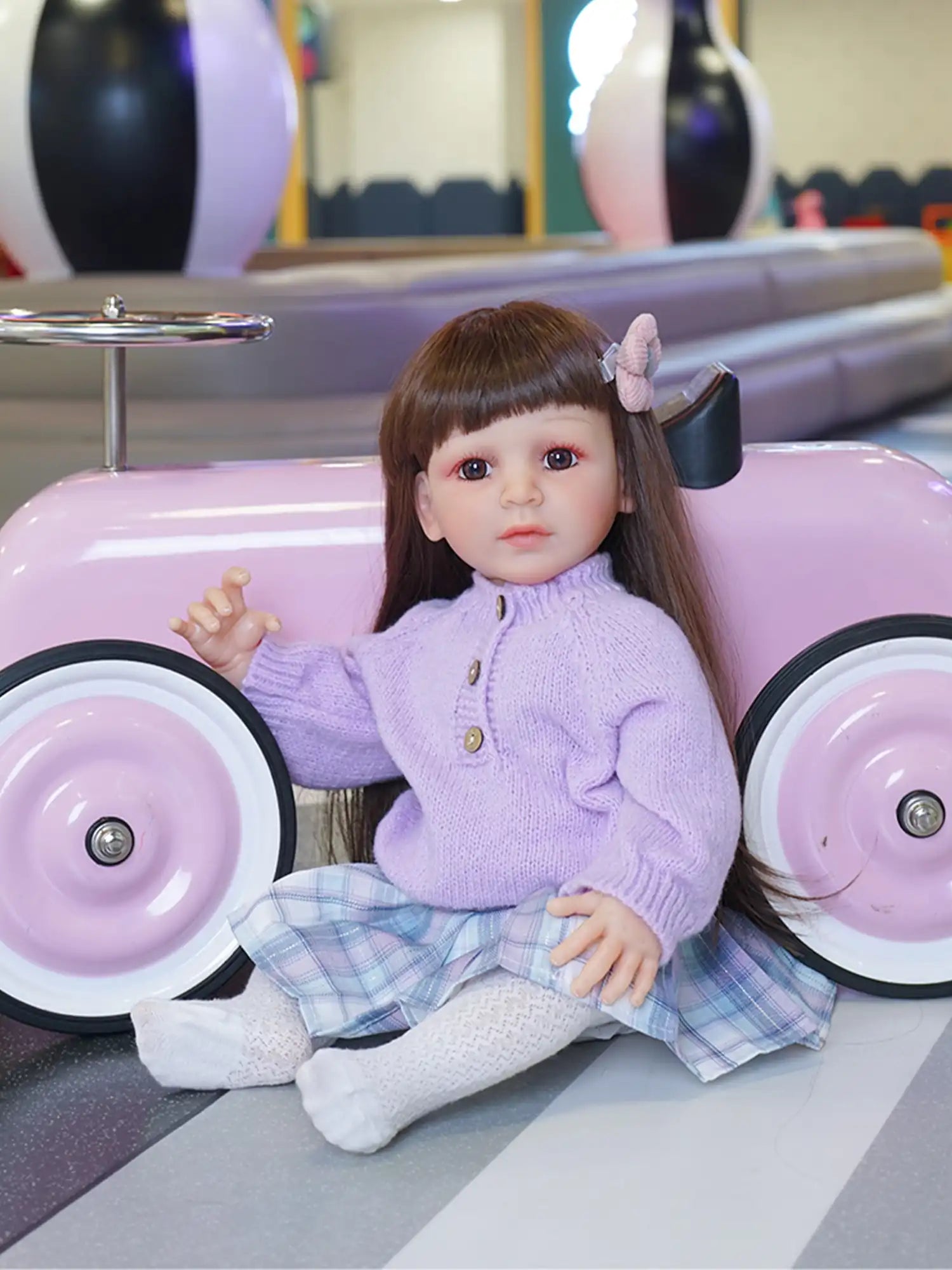 A toy toddler doll with a sweet face and brown hair with bangs, dressed in a pastel purple outfit, sitting by a toy car.