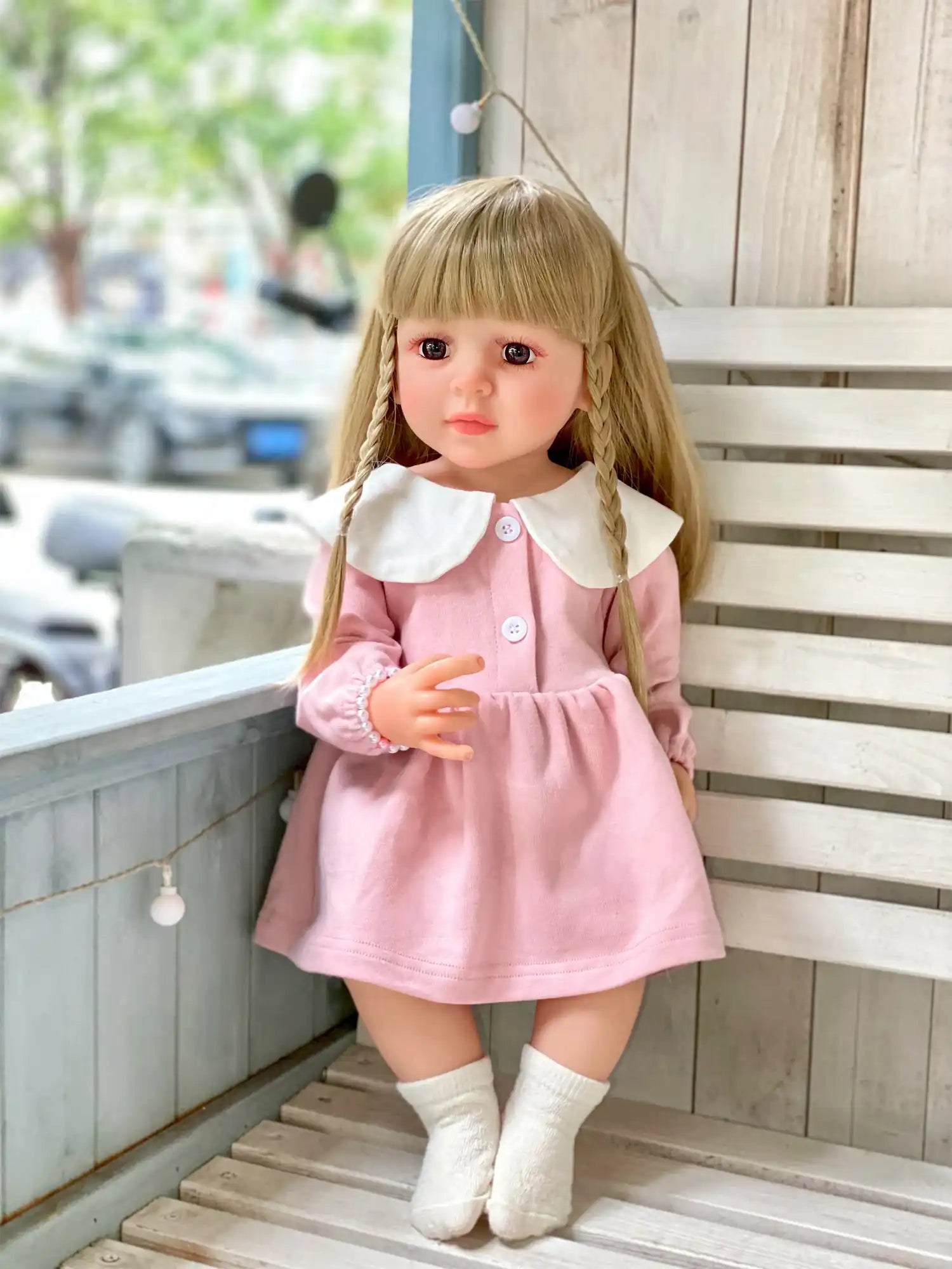 Realistic doll with braided hair and a collared pink dress, posed on white wooden steps.