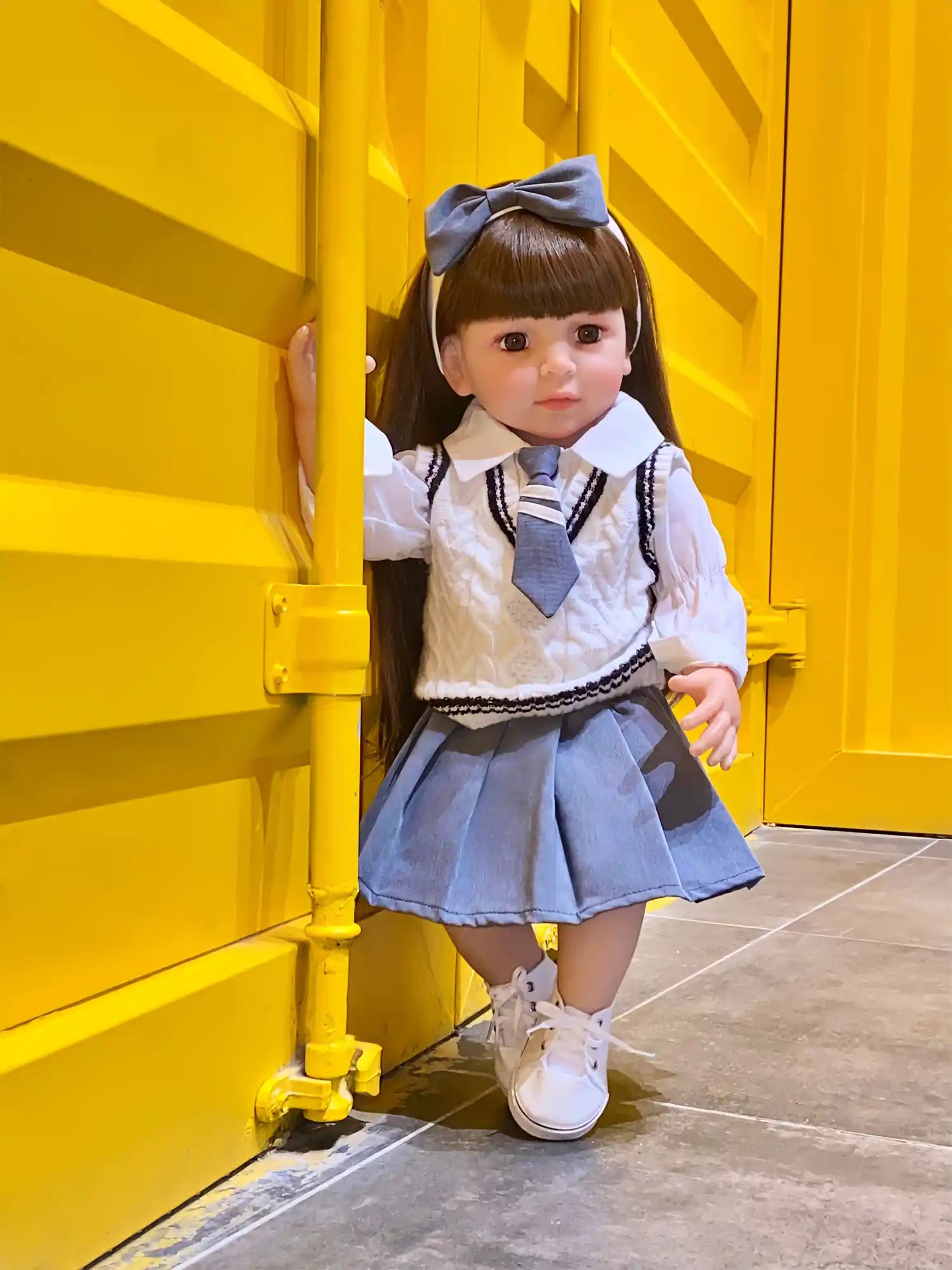 Lifelike toddler doll posed seated on a shiny floor, with a neatly styled hair, expressive eyes, and outfitted in a white blouse with a grey pinafore and a matching bow.