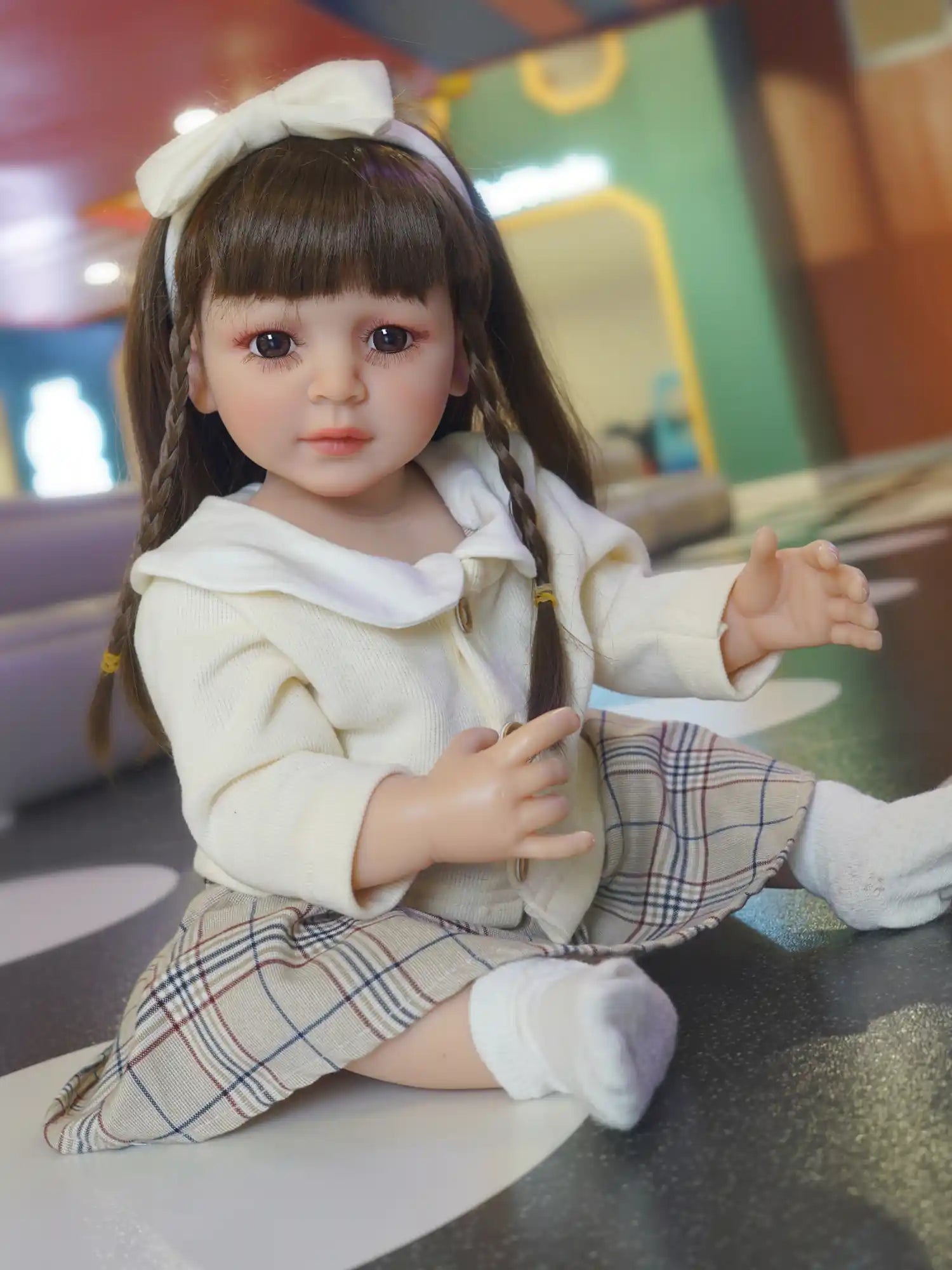 Realistic doll seated cross-legged with a gentle expression, in classic attire against a playful indoor backdrop.