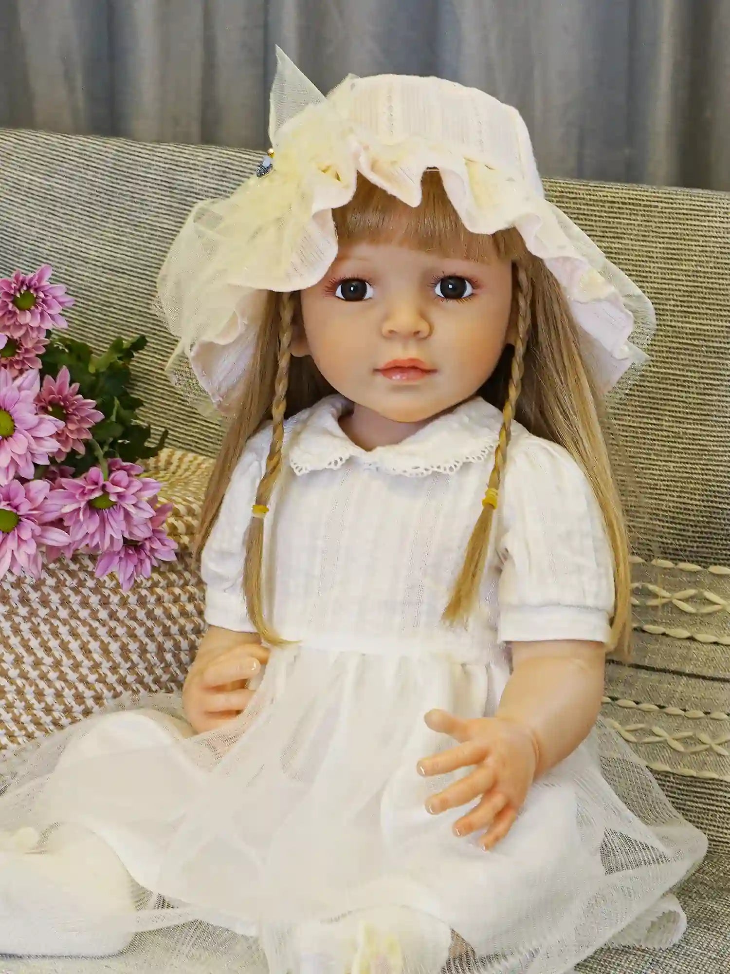 Realistic doll with long hair and a yellow bonnet, holding a flower, on a woven chair.