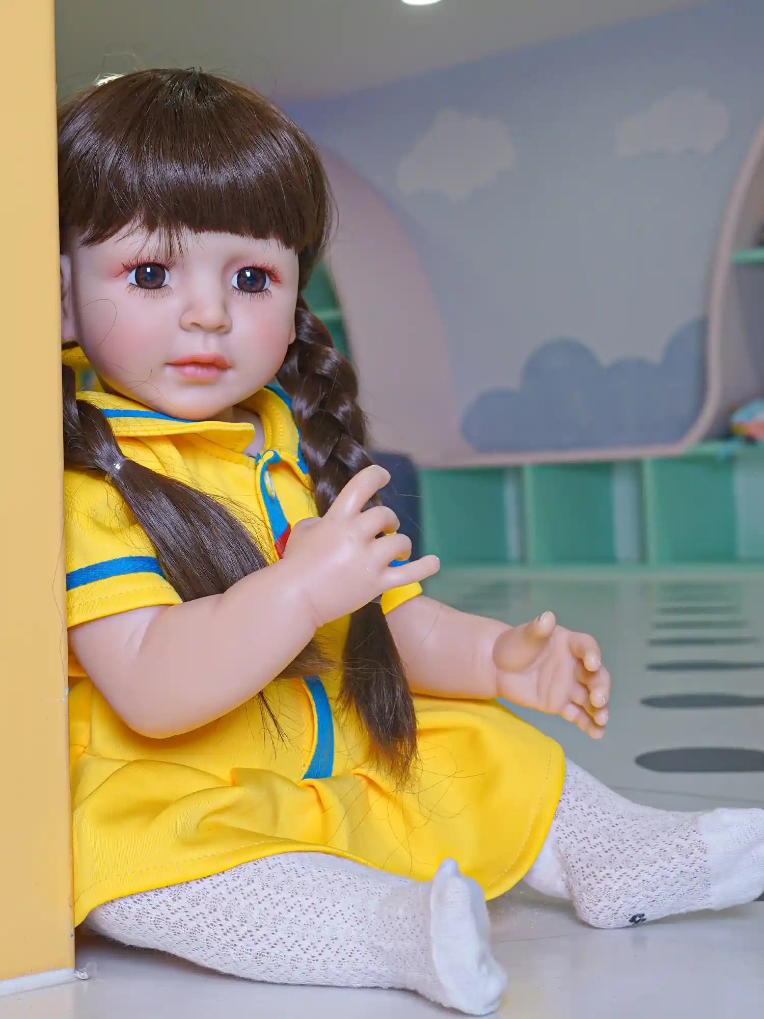 A lifelike doll, sporting a yellow dress with blue accents, sits casually against a yellow wall, complementing the colorful indoor playground.