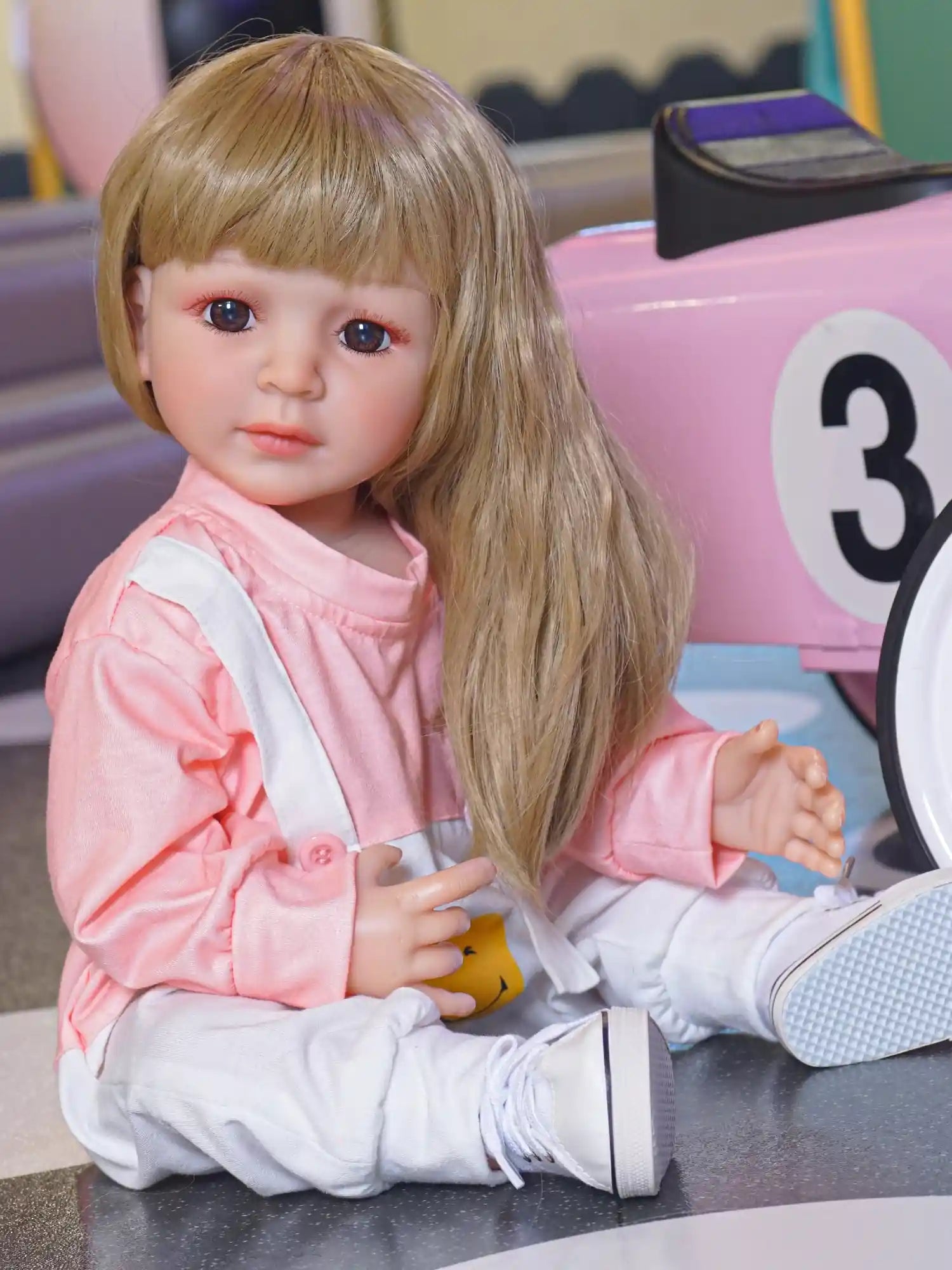 A realistic doll seated with a reflective expression, in a cozy pink and white outfit, beside a pink toy car.