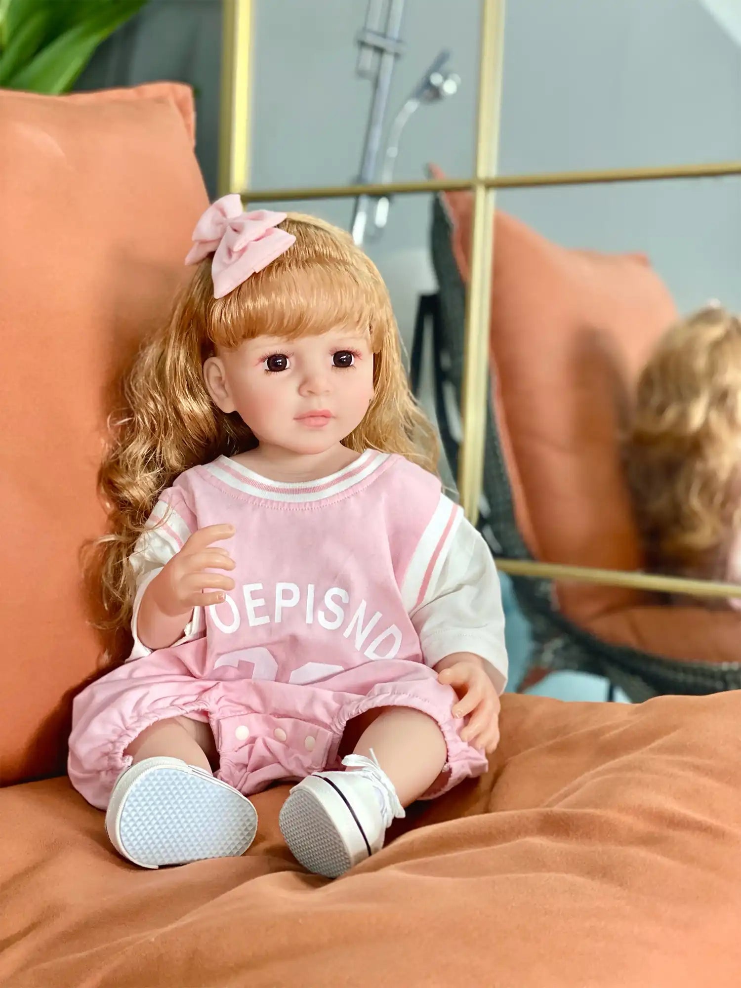 A strawberry blonde-haired doll with a pink bow, seated comfortably, wearing a pink jersey, shorts, and white sneakers.