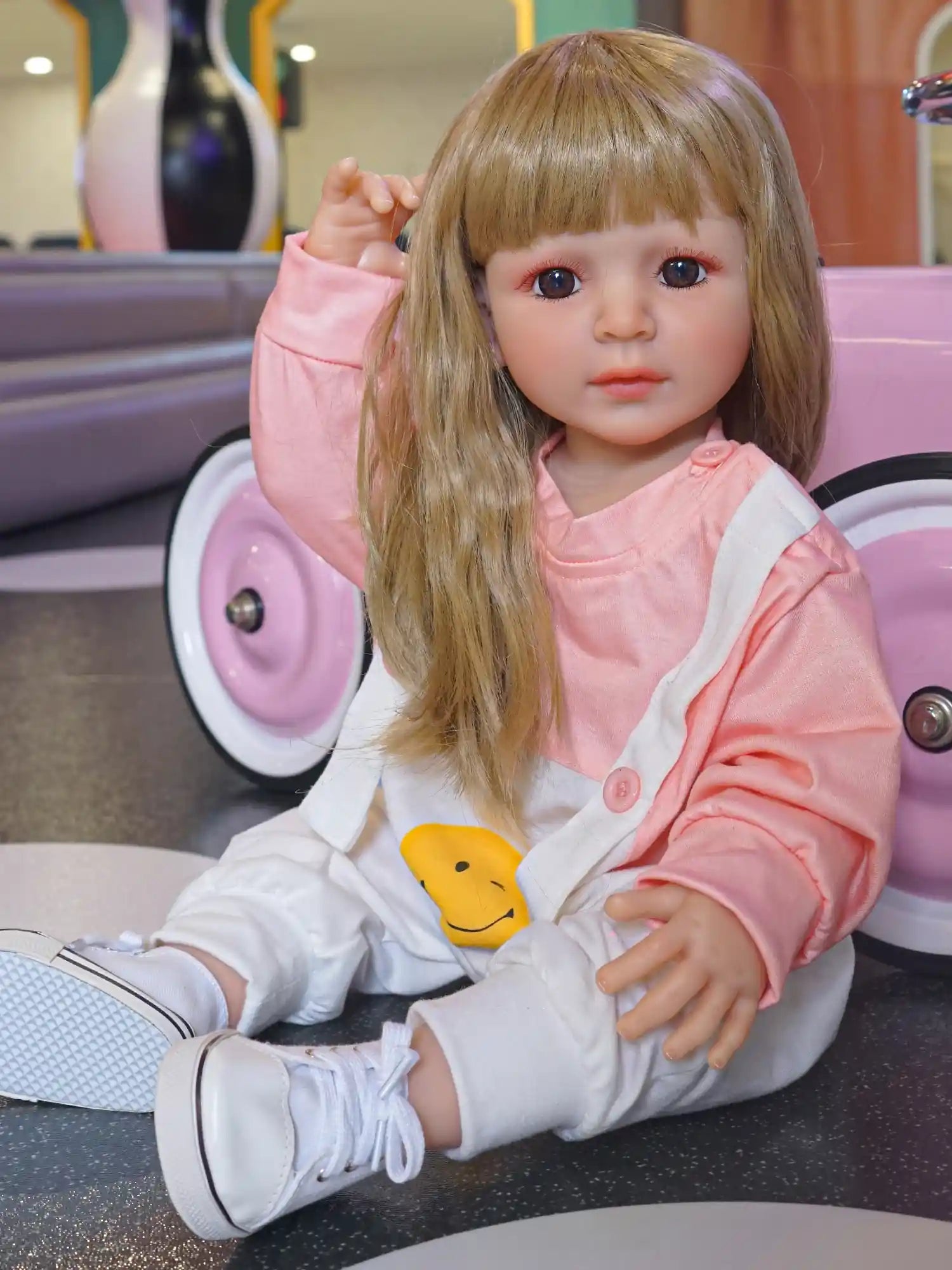Blonde-haired doll in a pink top, holding a bright yellow ball, seated on a gray floor with play equipment nearby.