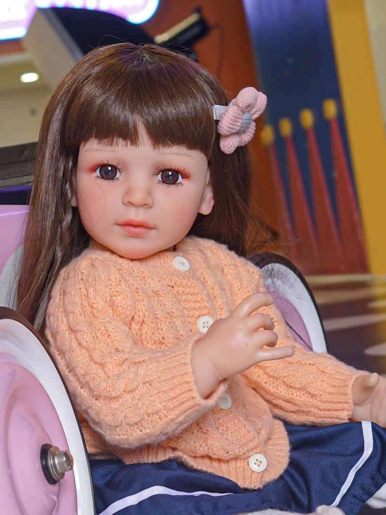 A realistic toddler doll, positioned seated with one hand raised, in casual attire against a playful backdrop.