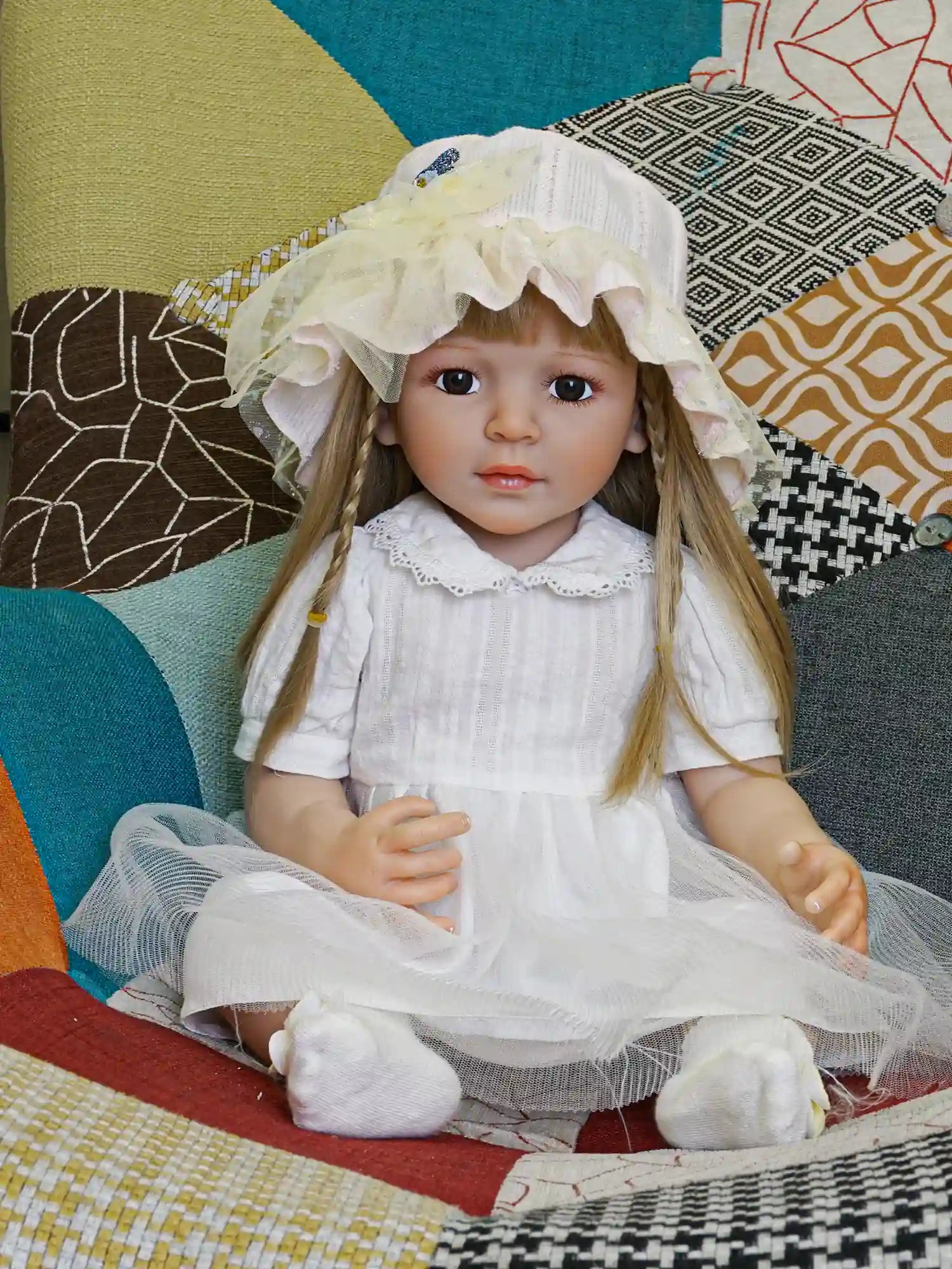 Toddler doll in white with a yellow hat, among pink flowers on a gray couch.