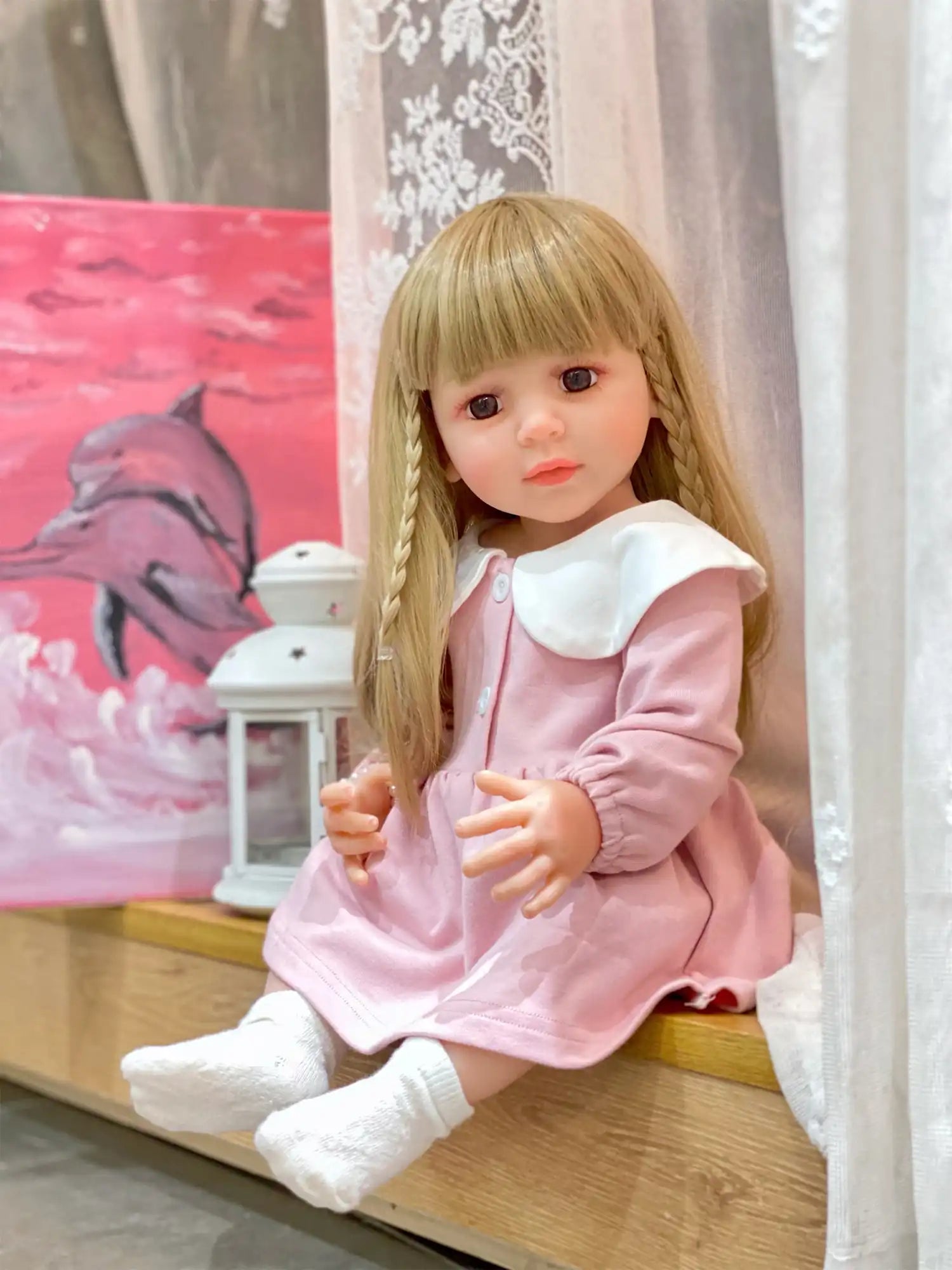 Doll with a contemplative look in a pastel pink dress, seated indoors with a dolphin painting in the background.