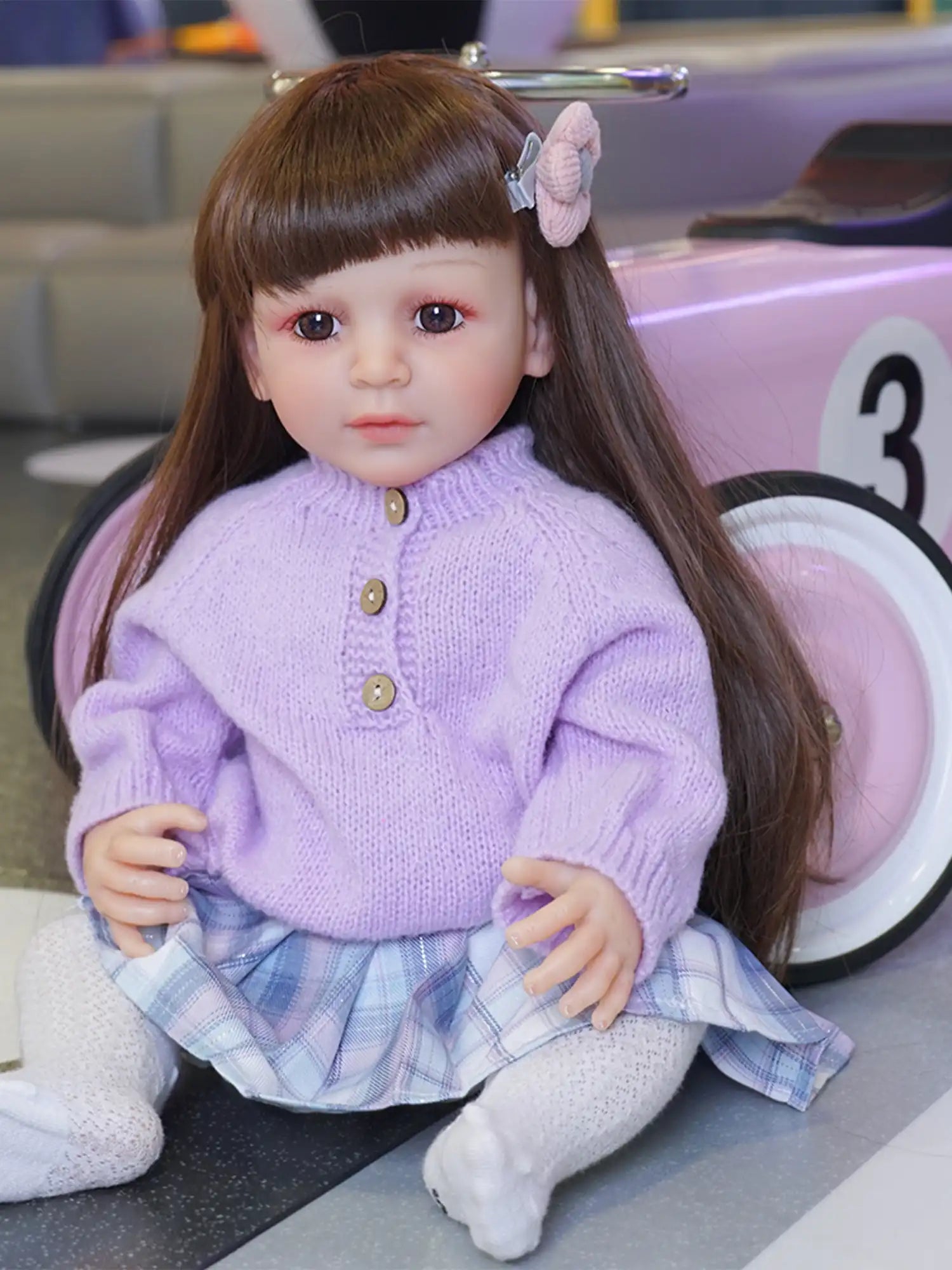 A lifelike toddler doll sits with a lilac knit sweater and a plaid skirt, her brown hair adorned with a pink bow clip.