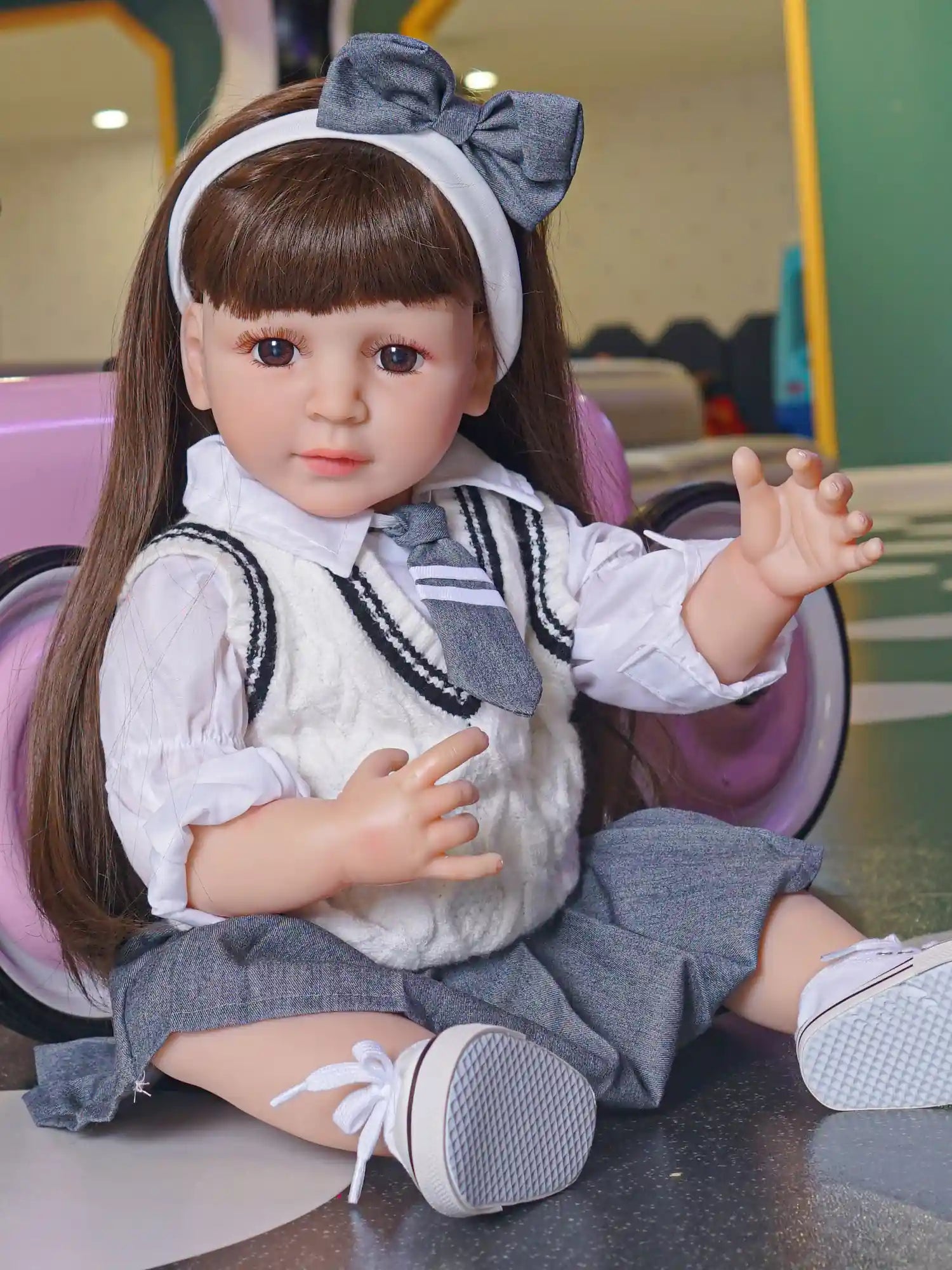 Detailed reborn doll with an attentive expression, dressed in a classic school uniform with a white blouse and grey skirt, and accessorized with a large bow headband.