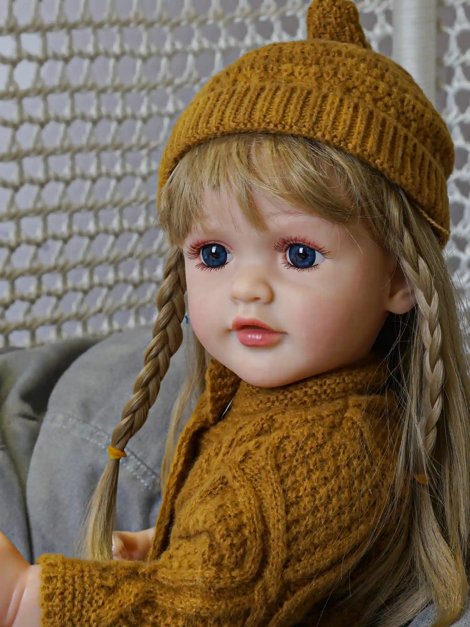 Doll designed to resemble a thoughtful child, with delicate facial features, a braid, and deep blue eyes, dressed warmly in a knitted mustard ensemble.