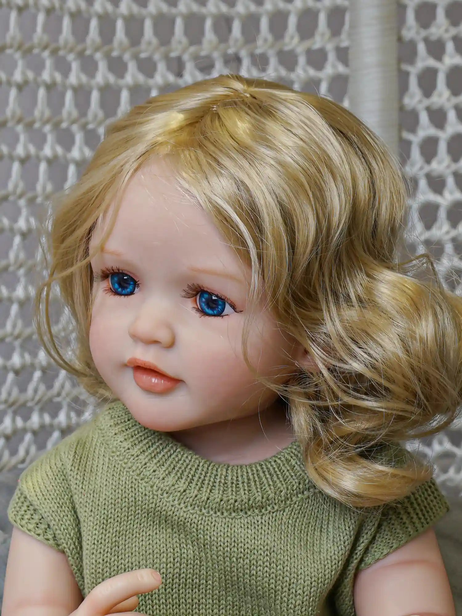 A lifelike reborn doll sitting comfortably, with soft blonde curls and clear blue eyes, dressed in a green outfit with a tulle skirt.