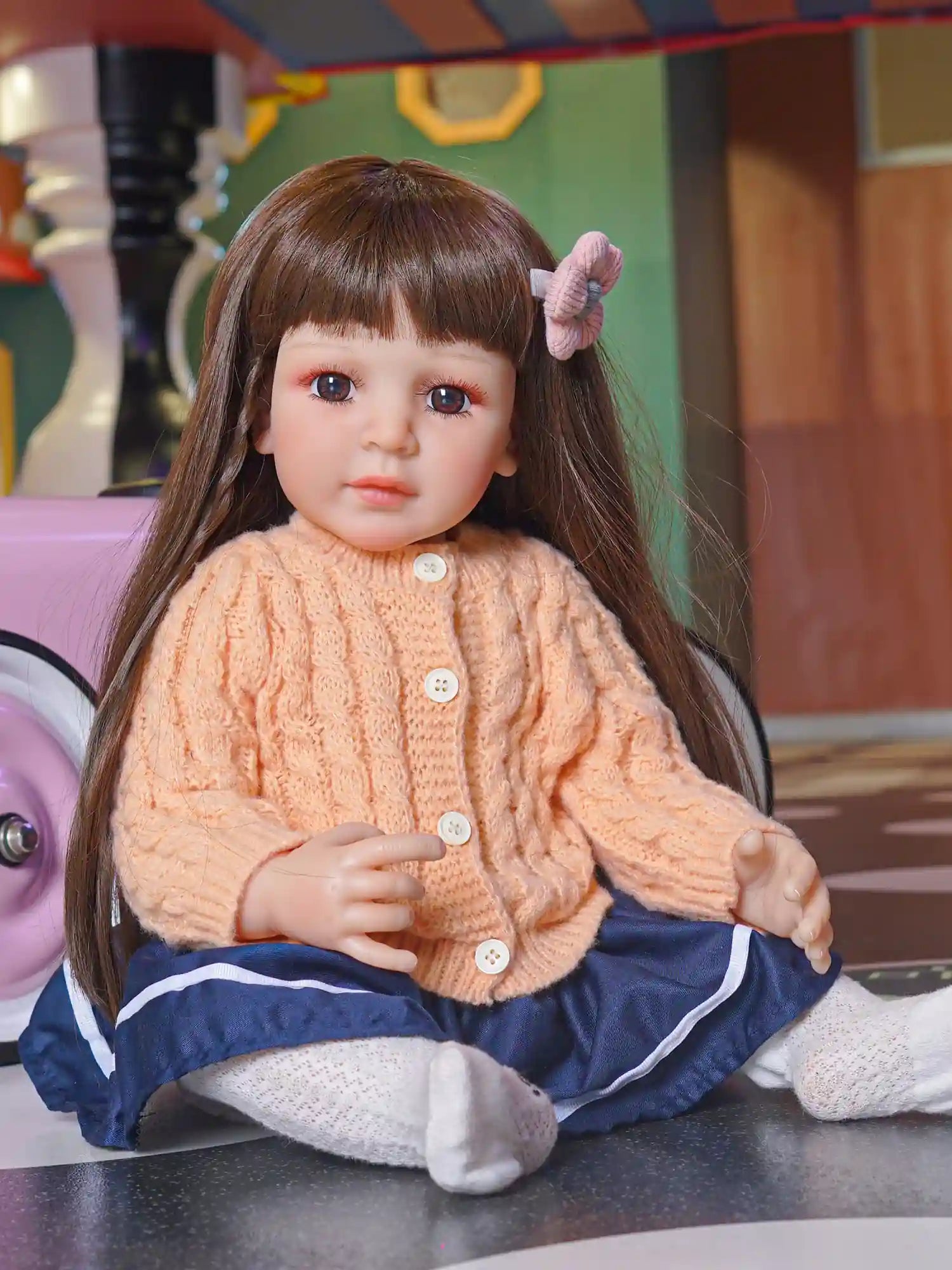 Toddler doll wearing a textured peach sweater and navy blue pants, with white lace tights, seated indoors.