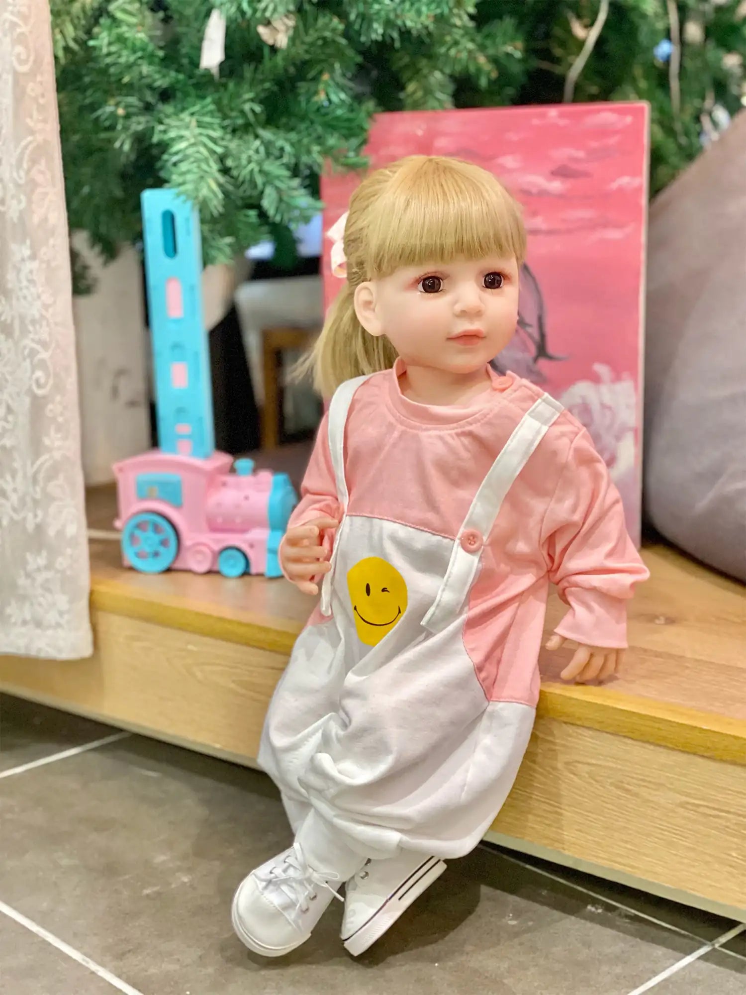 A doll with a delicate expression, dressed in a soft pink hoodie and white trousers, in a vibrant children’s play area.