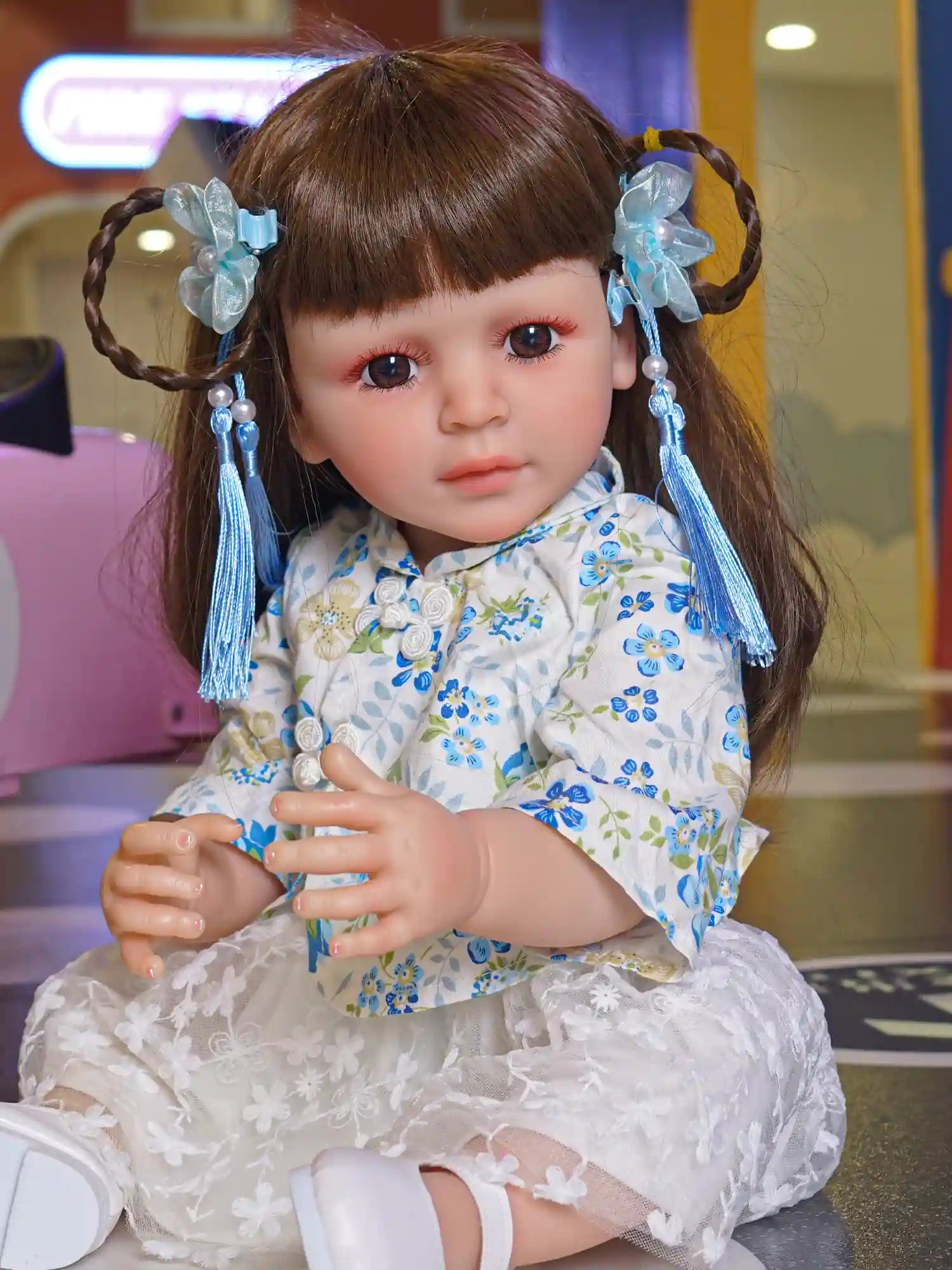 Toddler doll seated with hands clasped, wearing a blue floral dress and white shoes, with blue hair ribbons, in a playful indoor setting.