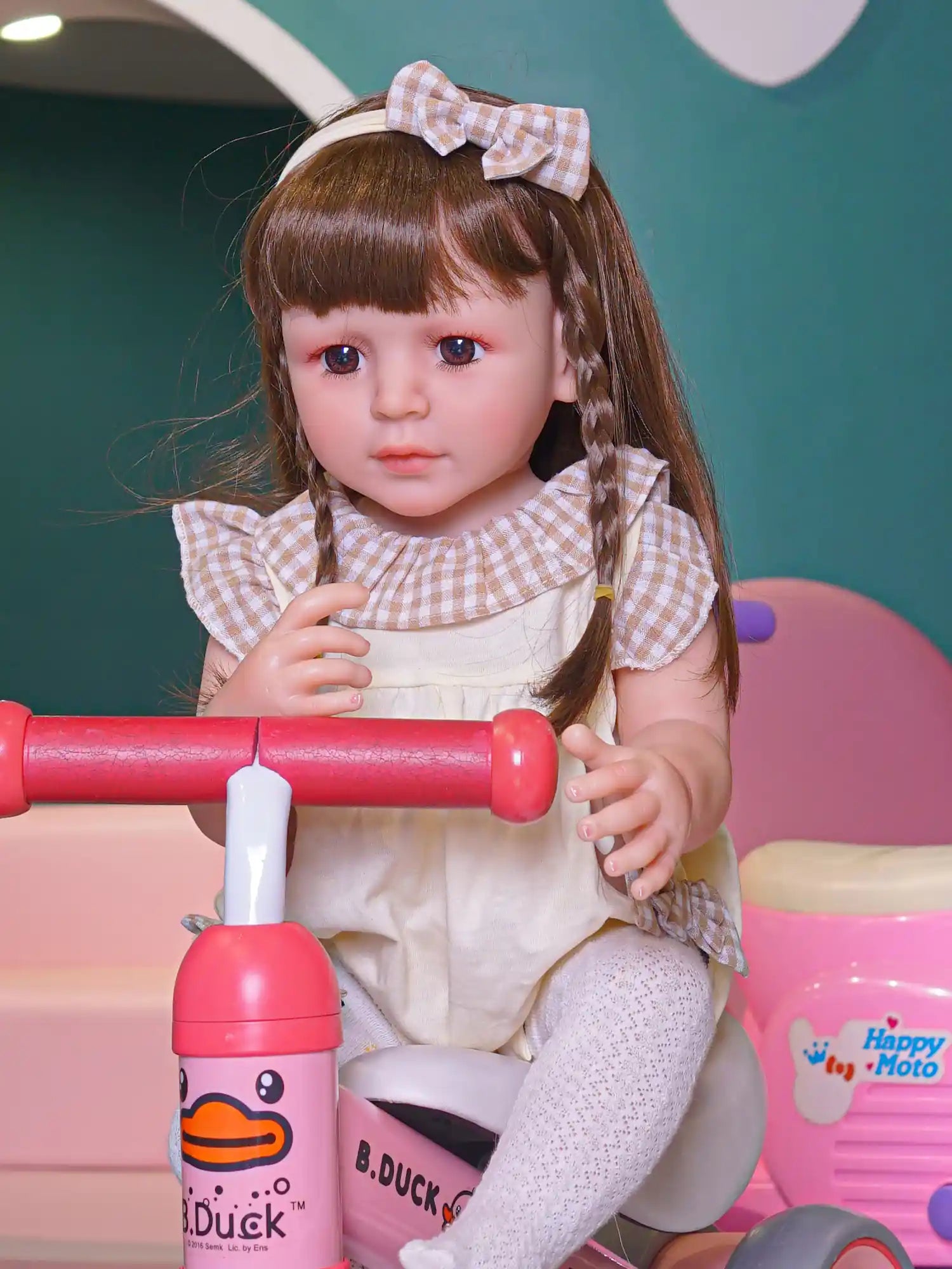 Realistic toddler doll dressed in a cream-colored outfit with white lace tights, seated indoors with toys.