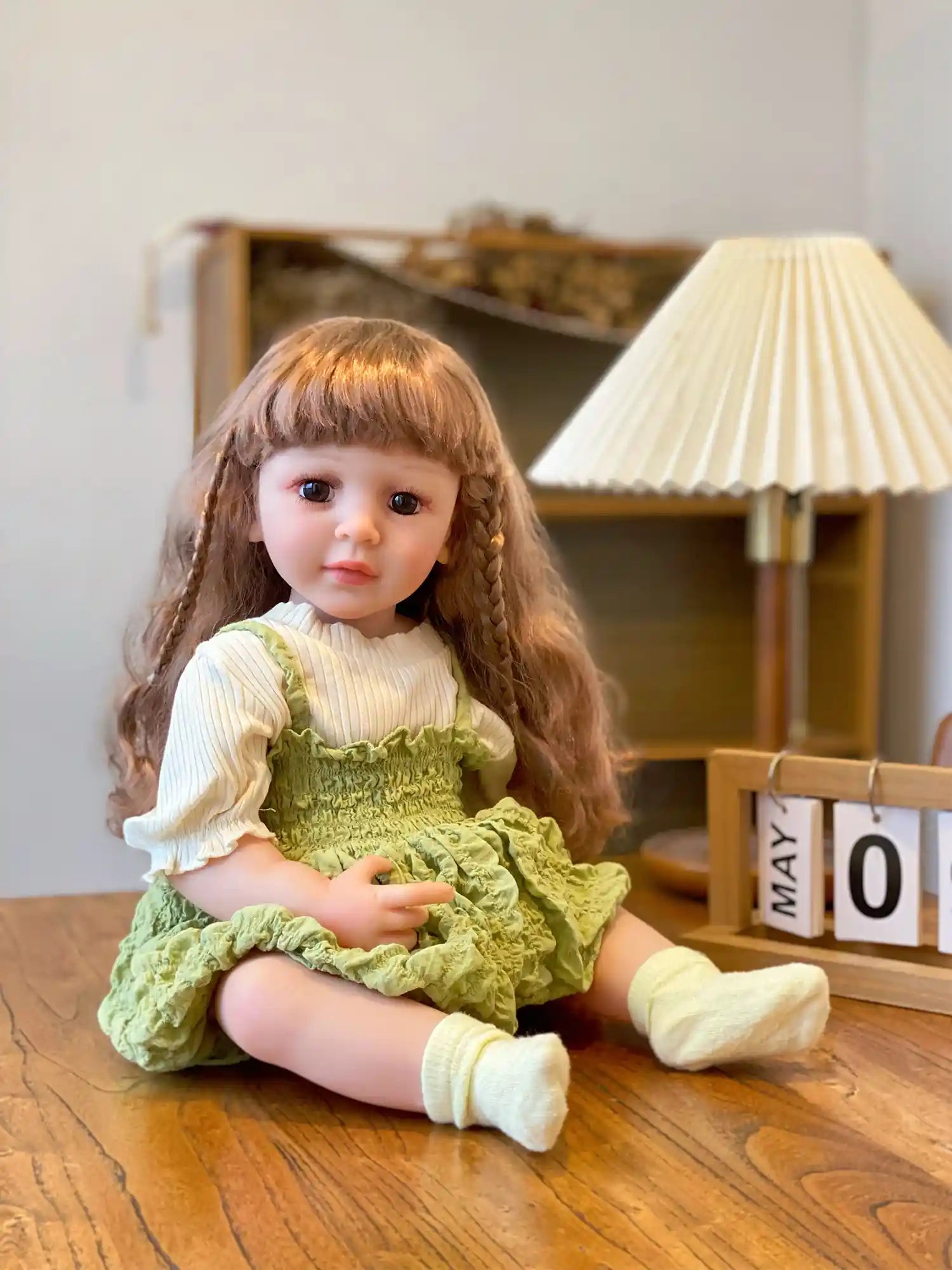 Realistic doll in green, sitting by a lamp and calendar on a wooden table.