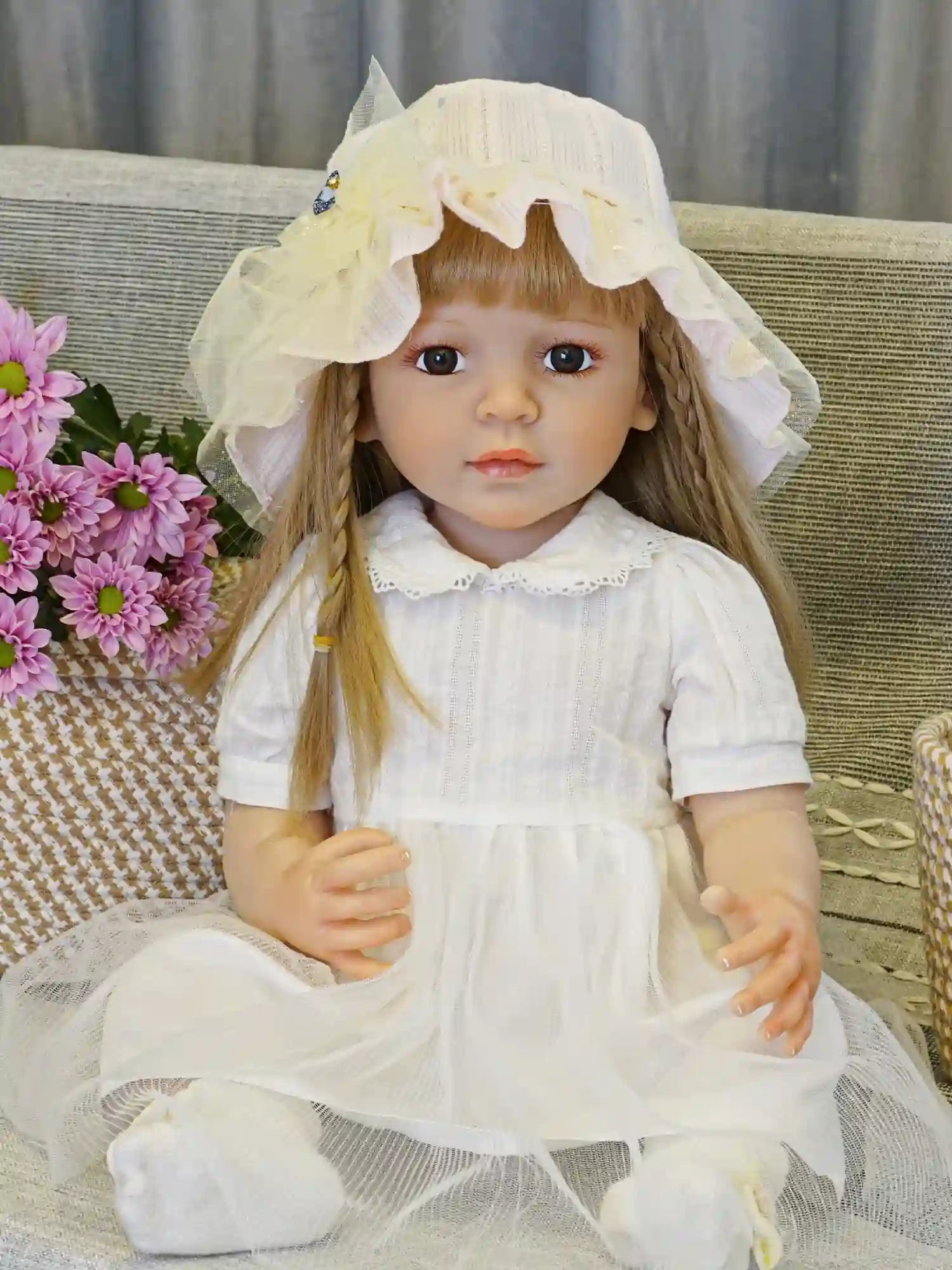 Collectible doll in a sunhat and white dress, seated, with a colorful cushion backdrop.