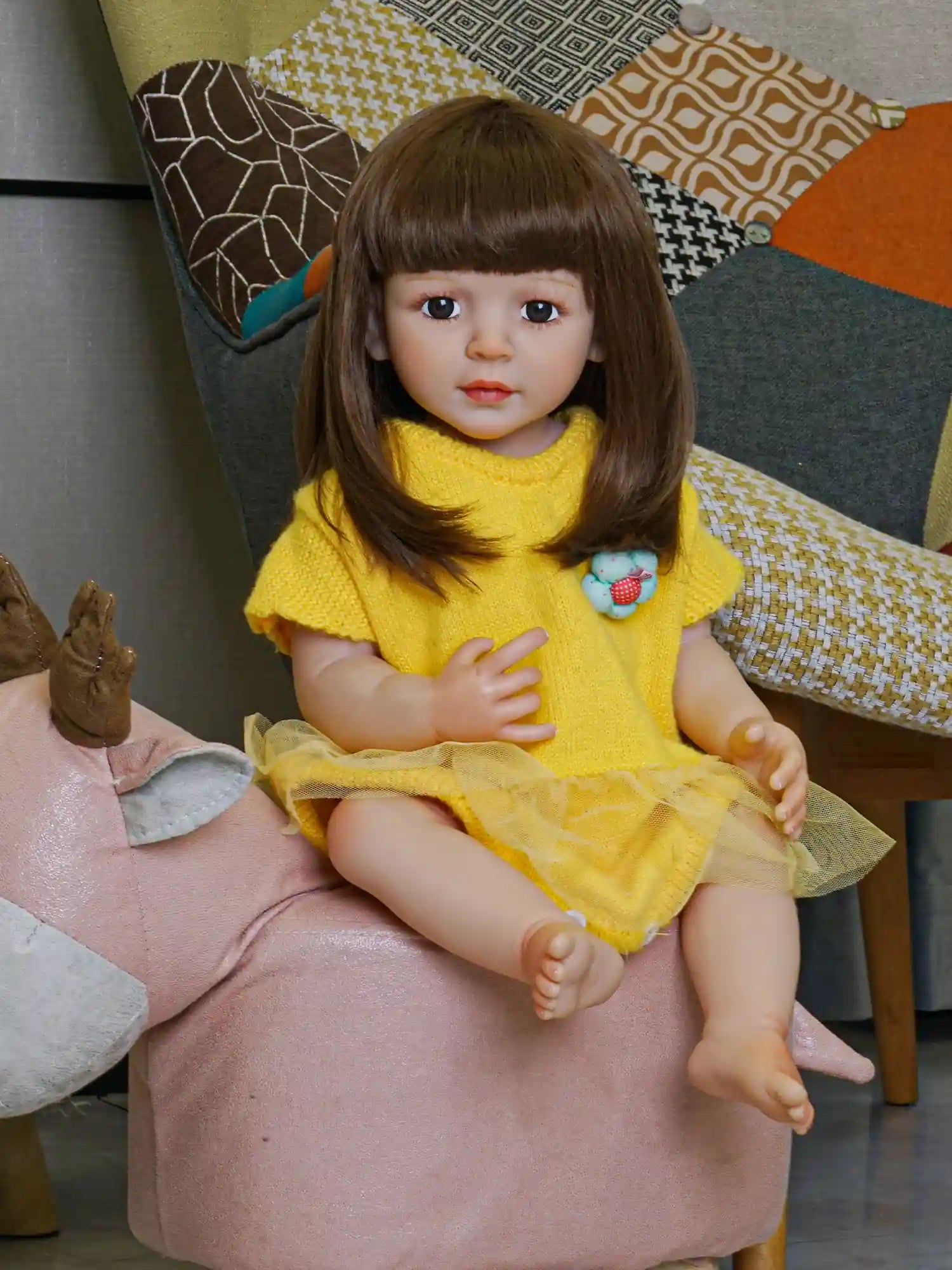 Realistic doll with a thoughtful gaze, clad in a cheerful yellow dress with a textured top and sheer overlay skirt, sitting against a modern geometric-patterned cushion.