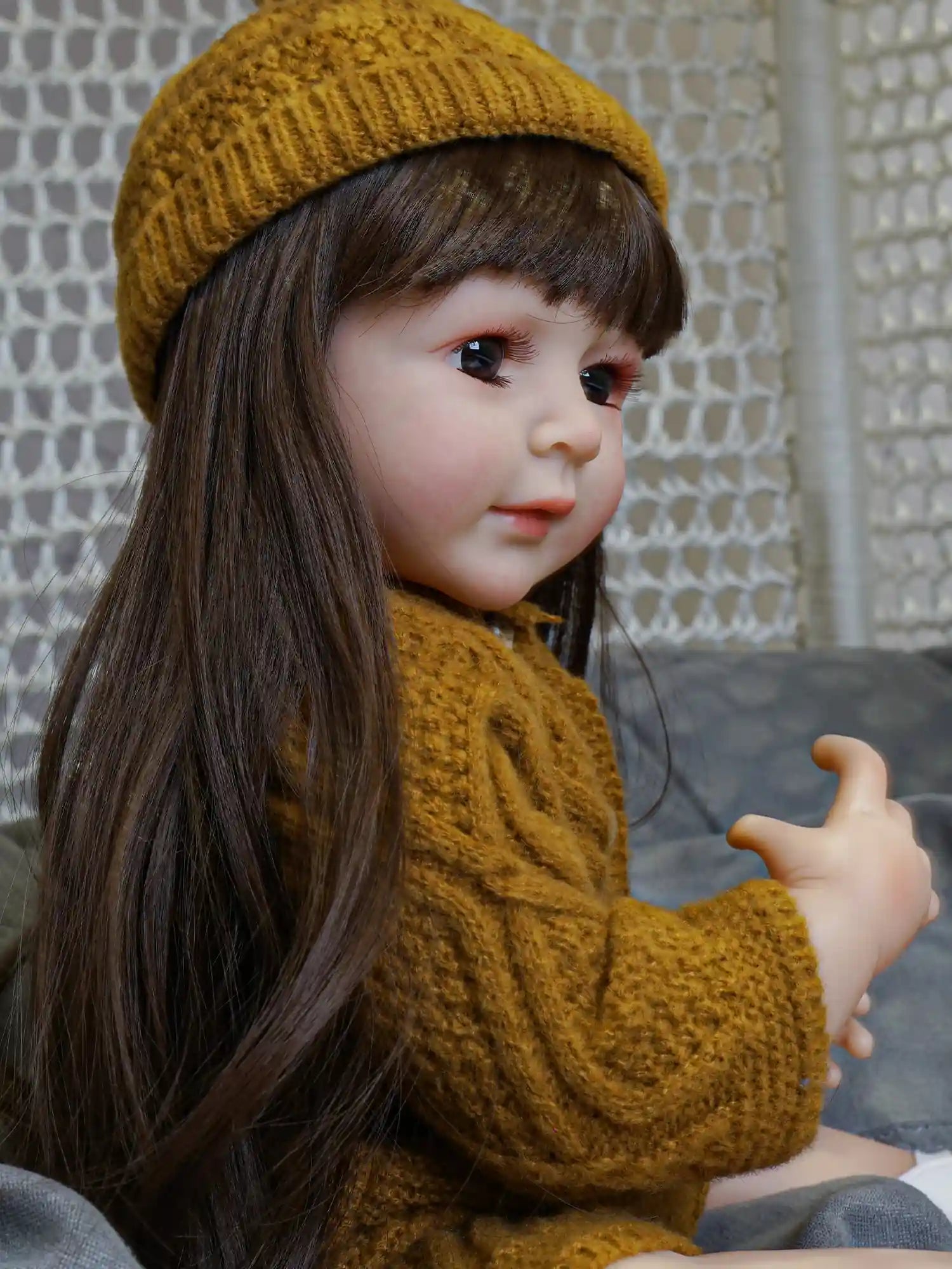 A reborn doll portraying a young girl, with deep brown eyes and a soft expression, wearing a knitted yellow hat and cardigan, set against a textured gray fabric.