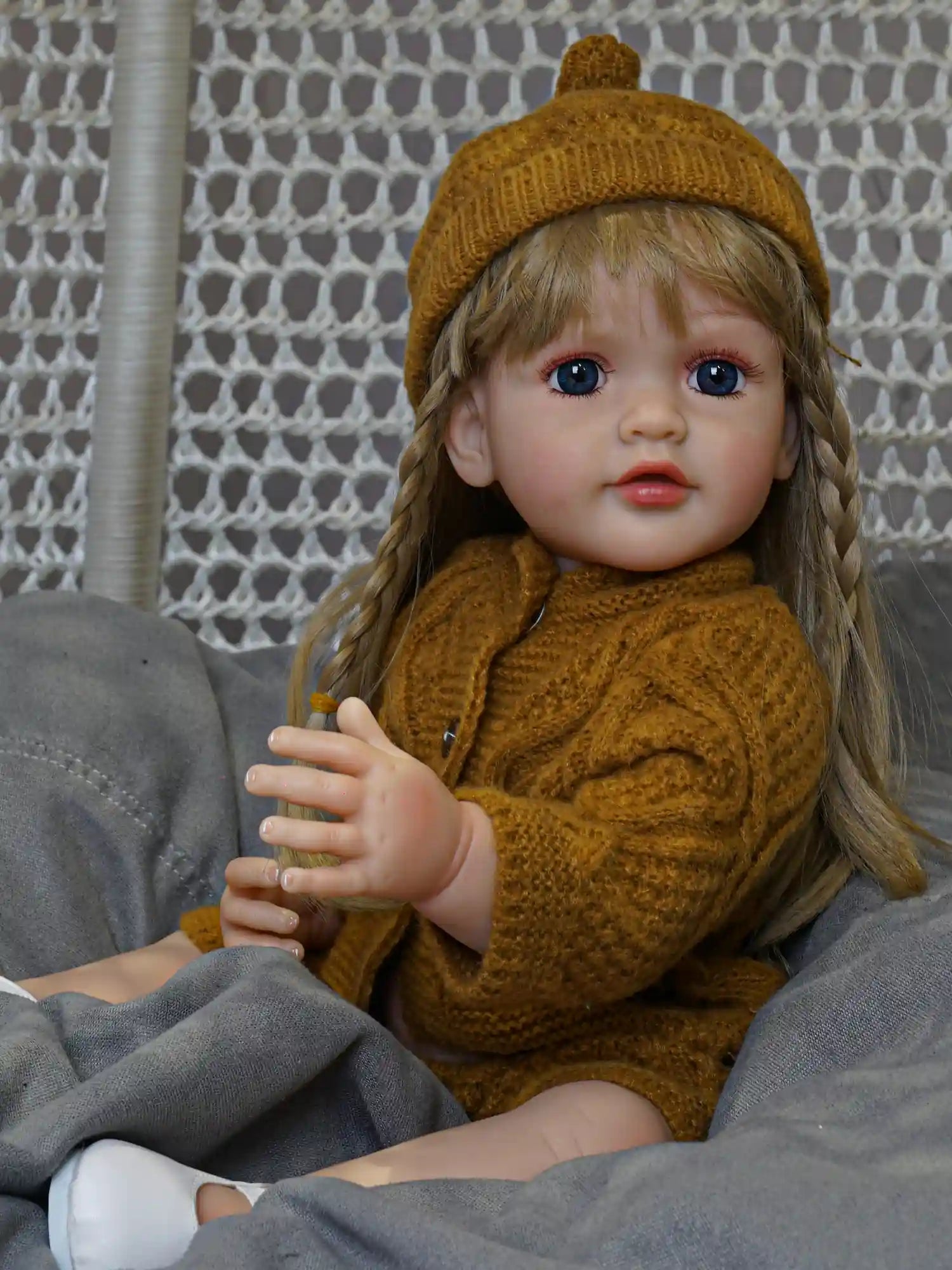 Seated reborn doll with blue eyes and a soft braided hairstyle, wearing a hand-knitted mustard sweater and cap, against a handcrafted grey backdrop.