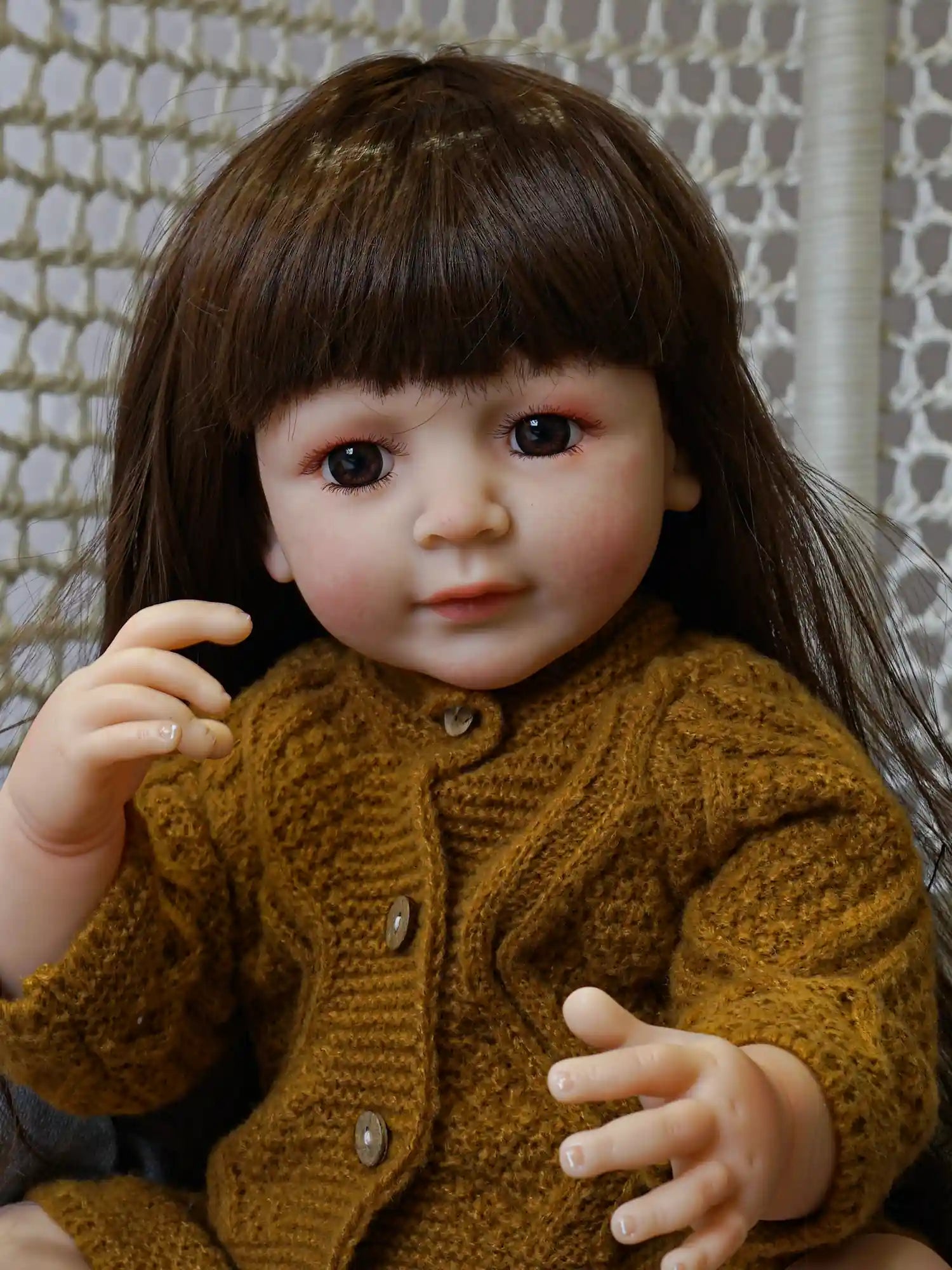 Artfully crafted doll resembling a seated toddler, with long brown hair under a yellow pompom hat, dressed in a chunky knit sweater in a warm mustard tone.