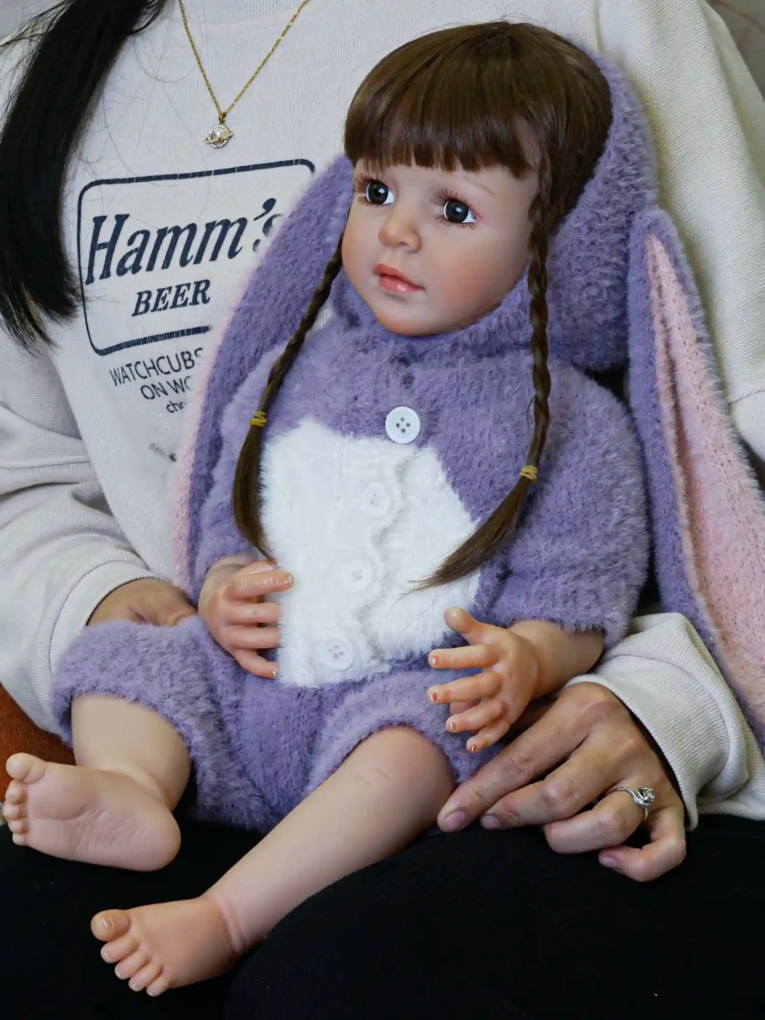 Toddler doll showcasing detailed craftsmanship, with shiny brown hair styled in braids and dressed in a purple bunny onesie, positioned against a neutral-toned backdrop.