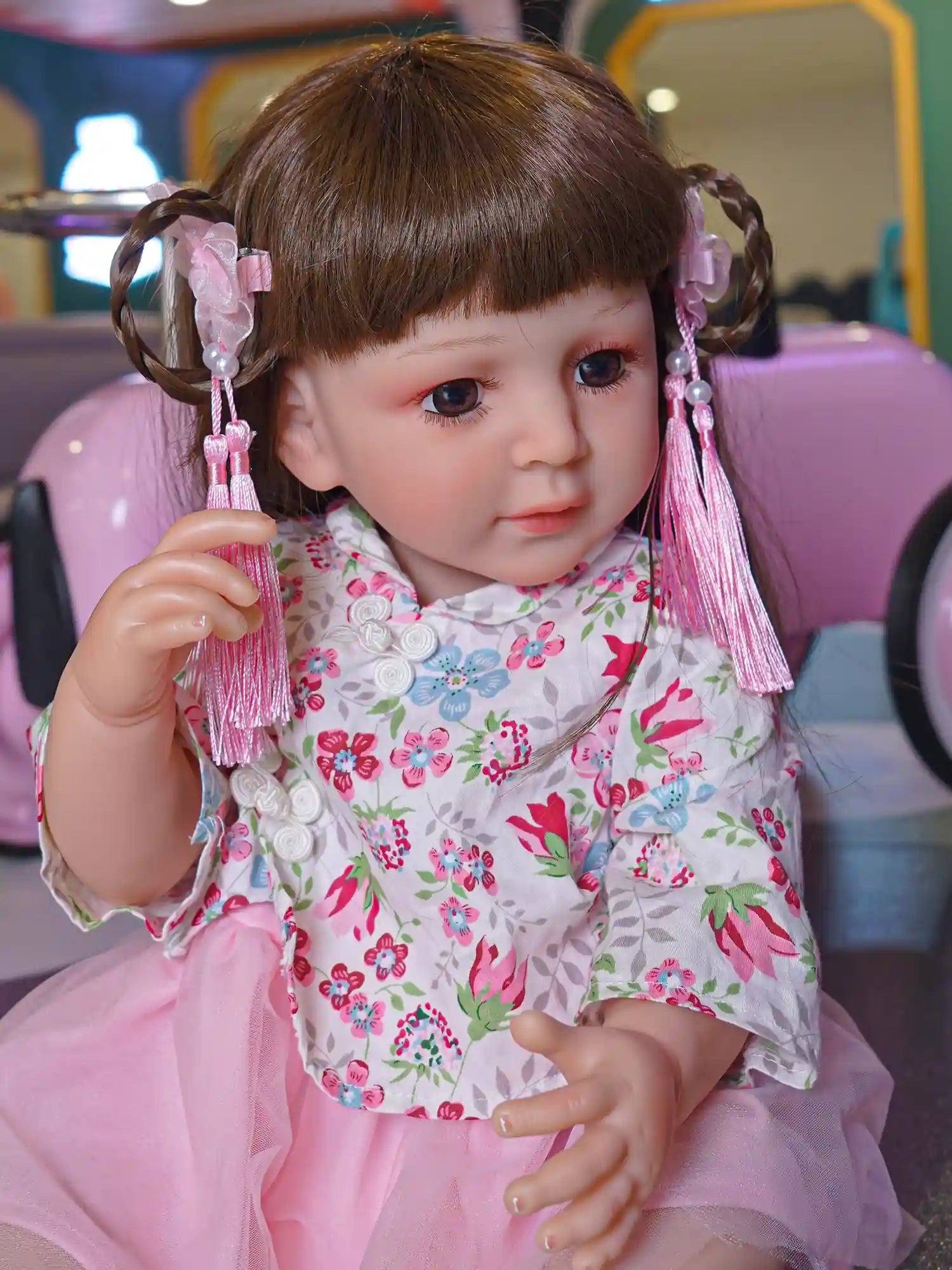 Toddler doll seated with legs crossed, dressed in a pink and white floral kimono dress with pink tulle skirt, white shoes, and pink ribbon hair accessories.