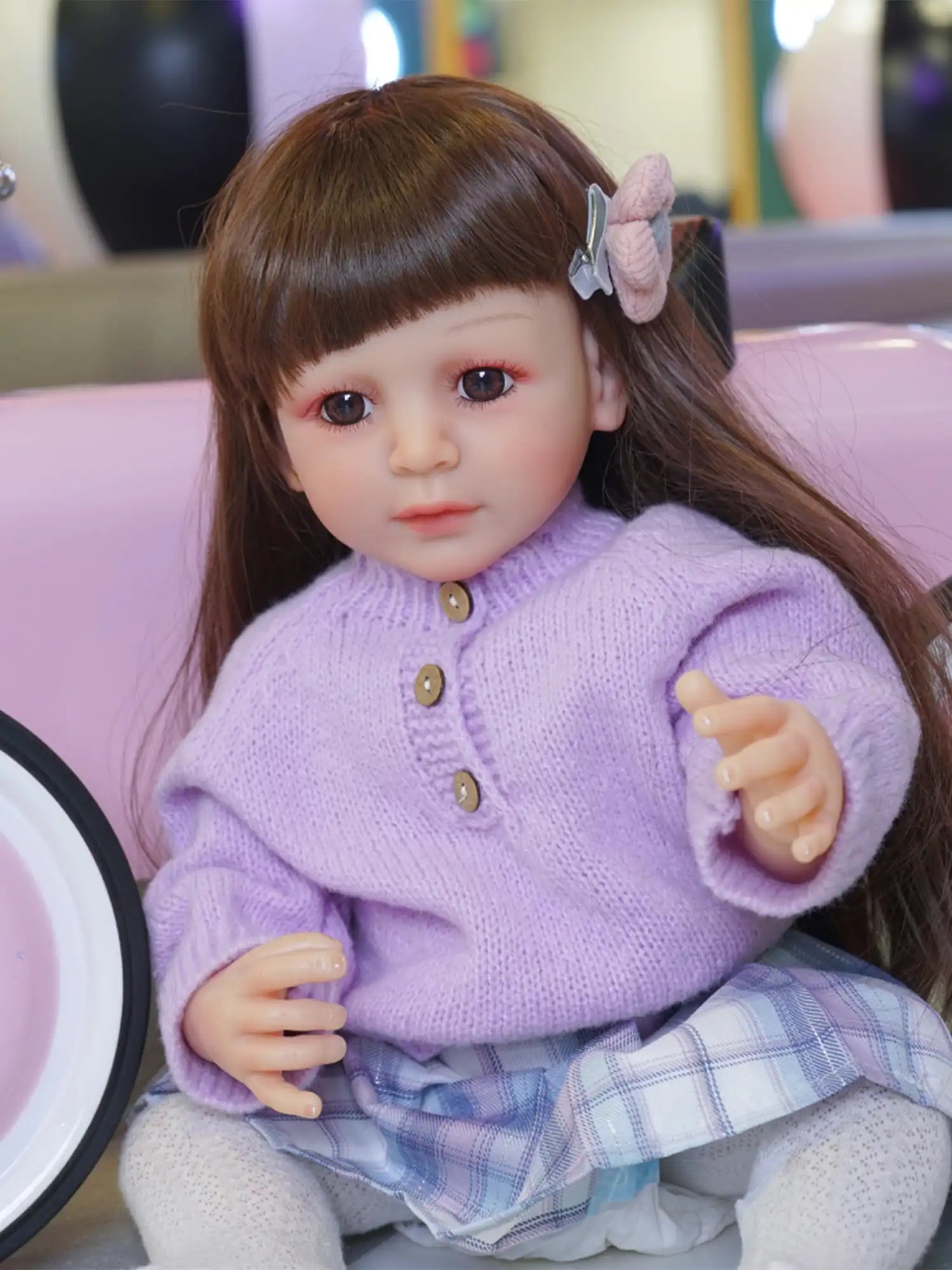 An adorable doll with lifelike features, wearing a textured purple sweater and a blue and white skirt, posing seated.