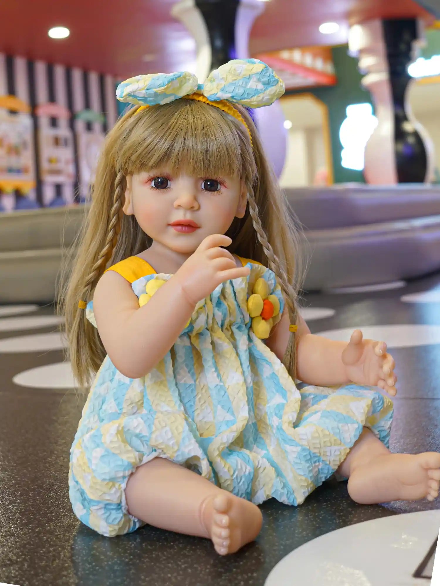 Realistic seated doll wearing a blue printed dress with yellow embellishments.