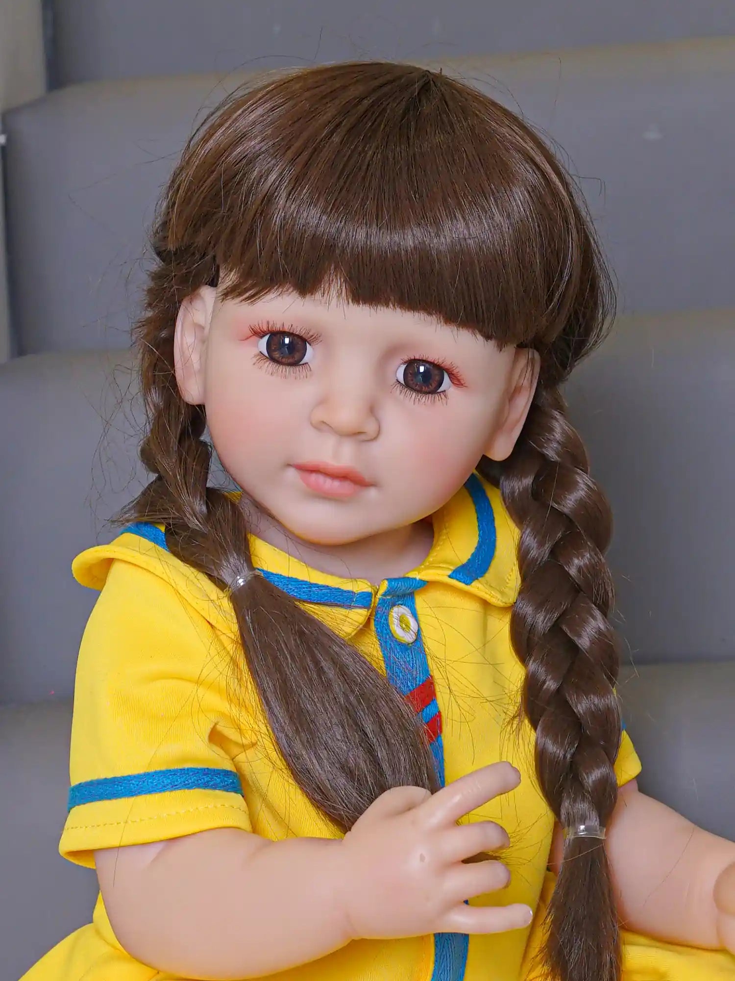 Realistic doll with braided locks and an observant expression, clad in a vibrant yellow outfit, inside a modern playroom.