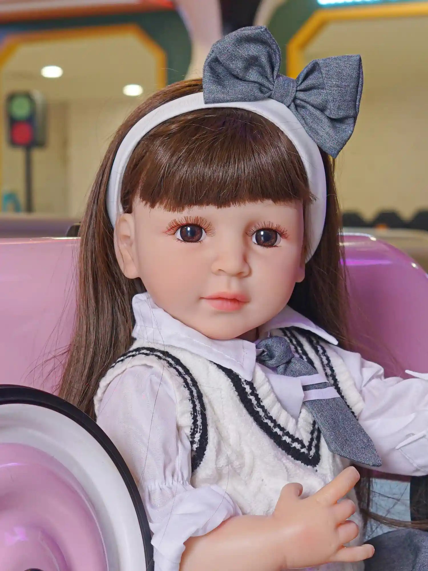 A collectible doll that resembles a little schoolgirl, with brown hair and engaging eyes, dressed in a white blouse and grey jumper, raising her hand as if to speak.