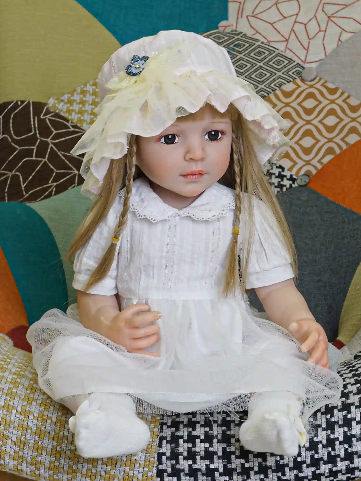 Lifelike reborn doll with light brown braided hair, wearing a white vintage-style dress and a delicate yellow sunhat, sitting on a grey armchair.