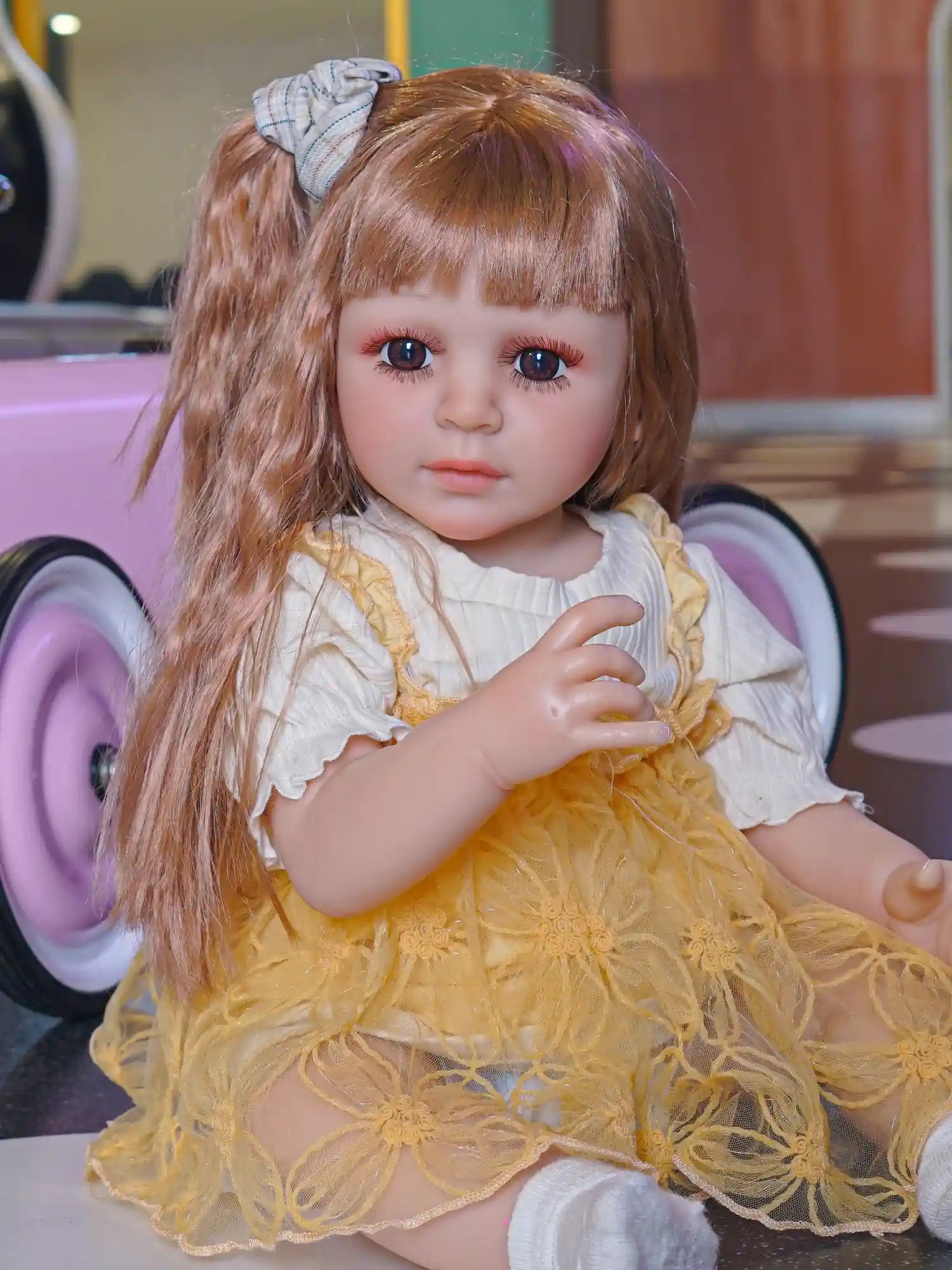 Lifelike doll showcasing large blue eyes, styled in a mustard-colored dress with detailed embroidery, in a playful setting.