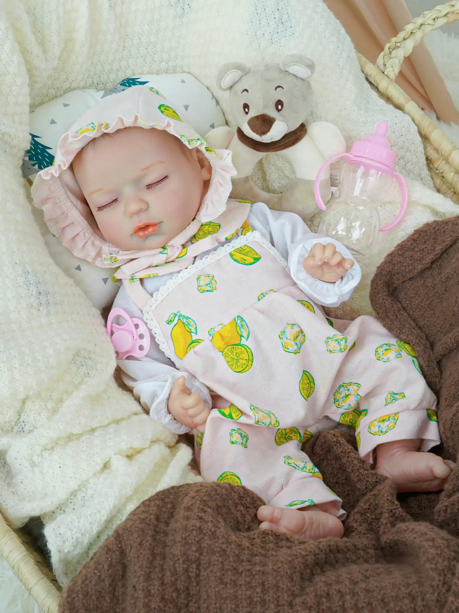 A reborn baby doll dressed in a pink bonnet with lemon prints and a matching outfit sleeps peacefully in a basket, surrounded by soft white blankets, with a plush teddy bear and a pink baby bottle nearby.