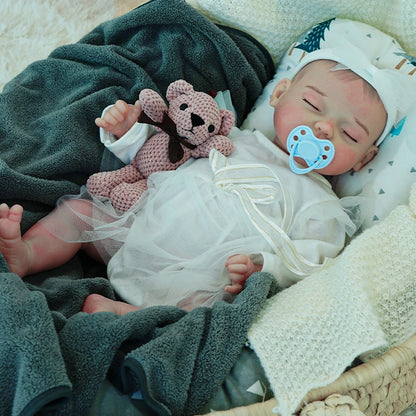 A reborn baby doll with a pacifier, dressed in a white outfit, lying on a dark blanket, holding a pink teddy bear.