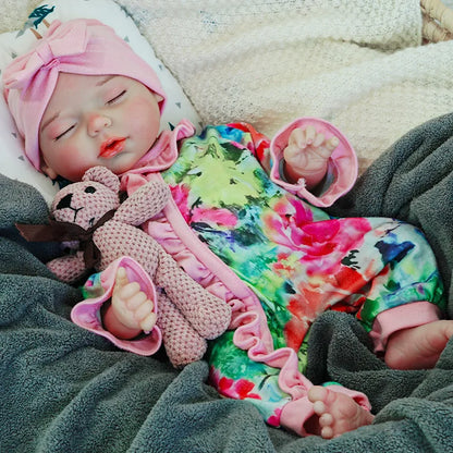 A reborn baby doll dressed in a colorful outfit with a pink headband, sleeping peacefully on a dark blanket, holding a pink teddy bear.